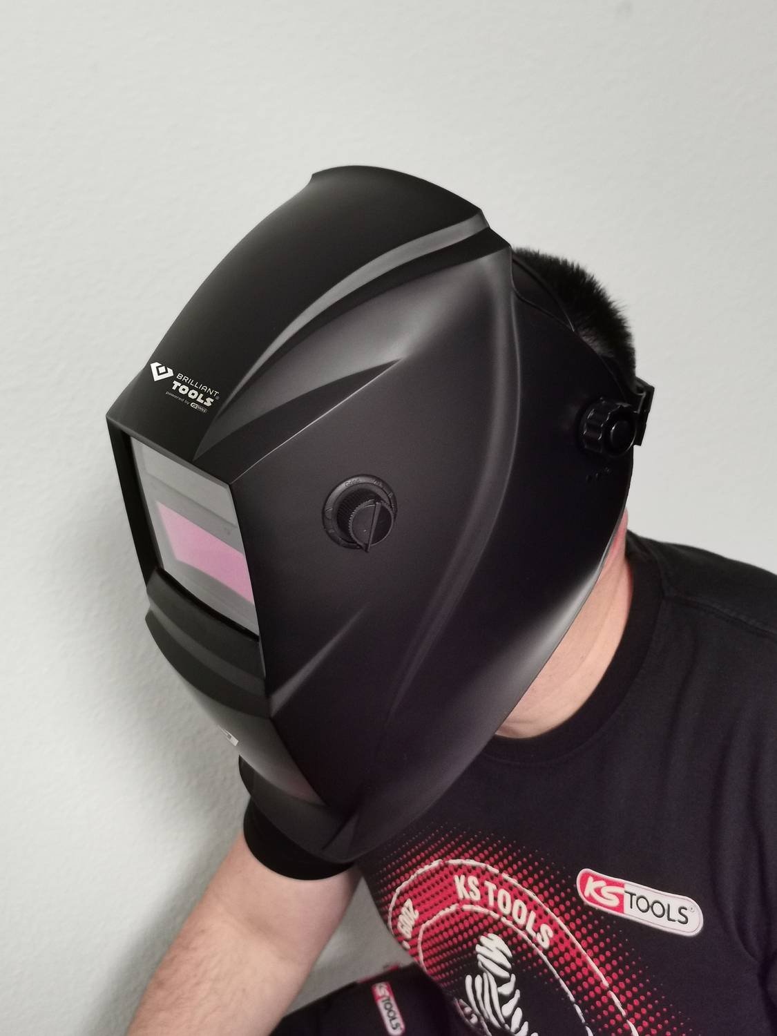 Osoba nosi czarny kask spawalniczy. Przyłbica jest lekko zabarwiona na różowo. Osoba ma na sobie czarny t-shirt z logo marki.