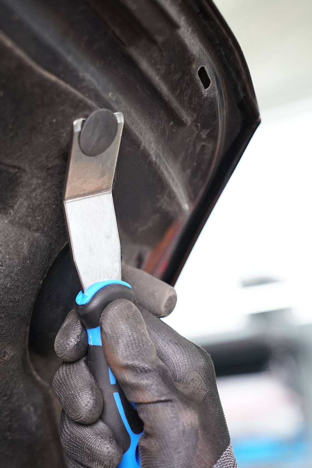 Ręka w rękawicy trzyma plastikowe narzędzie do pielęgnacji samochodu przy drzwiach pojazdu. Narzędzie jest wsuwane pod tapicerkę wewnętrzną.