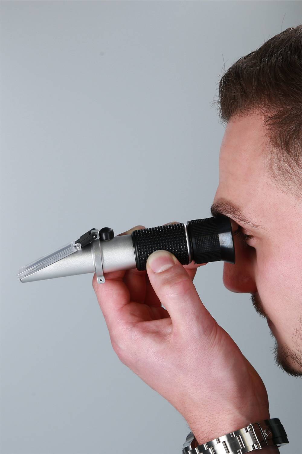 Mężczyzna patrzy przez srebrny refraktometr, który trzyma w prawej ręce.