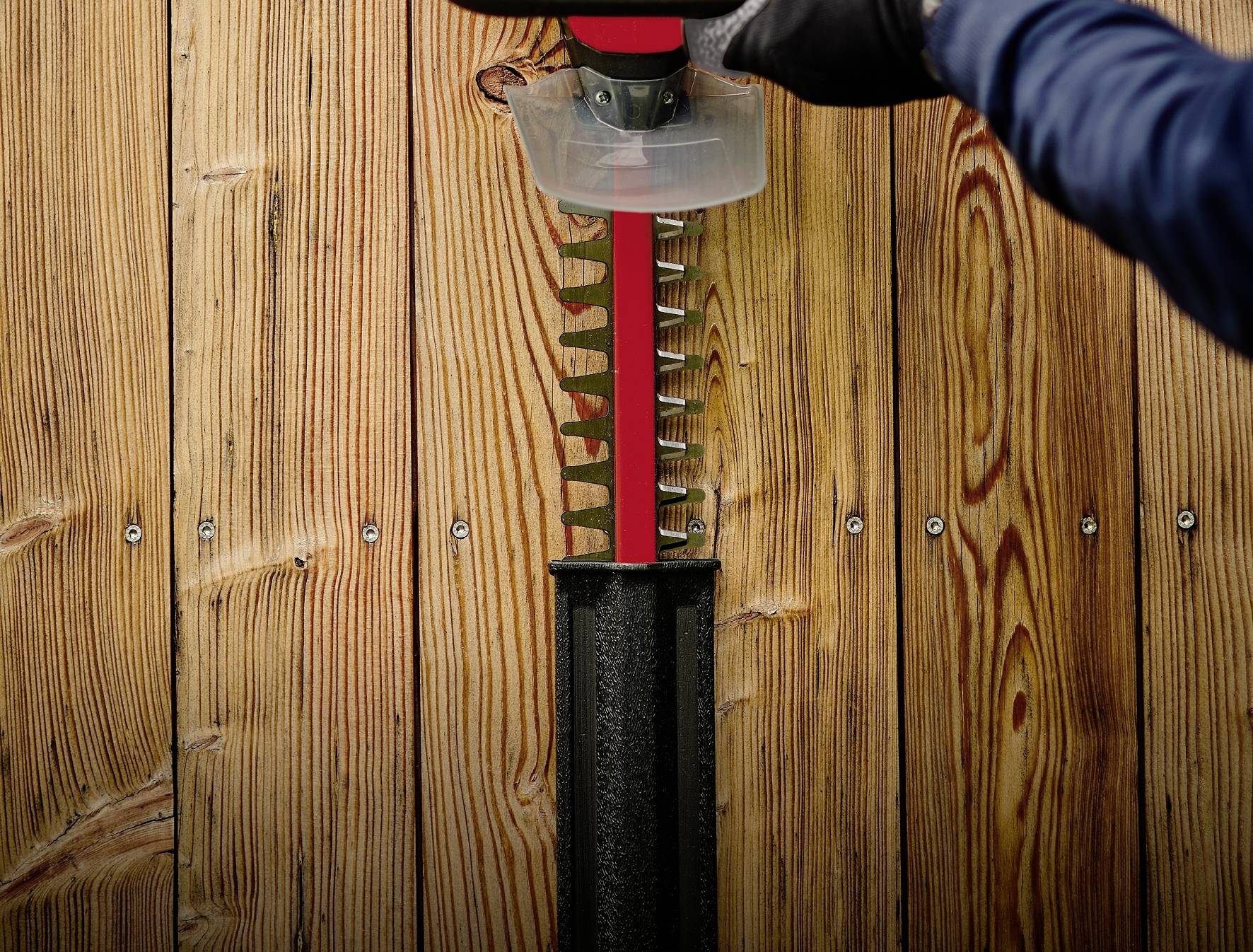 Osoba ostrzącą nożyce do żywopłotu za pomocą narzędzia do ostrzenia. W tle widoczna jest drewniana ściana.