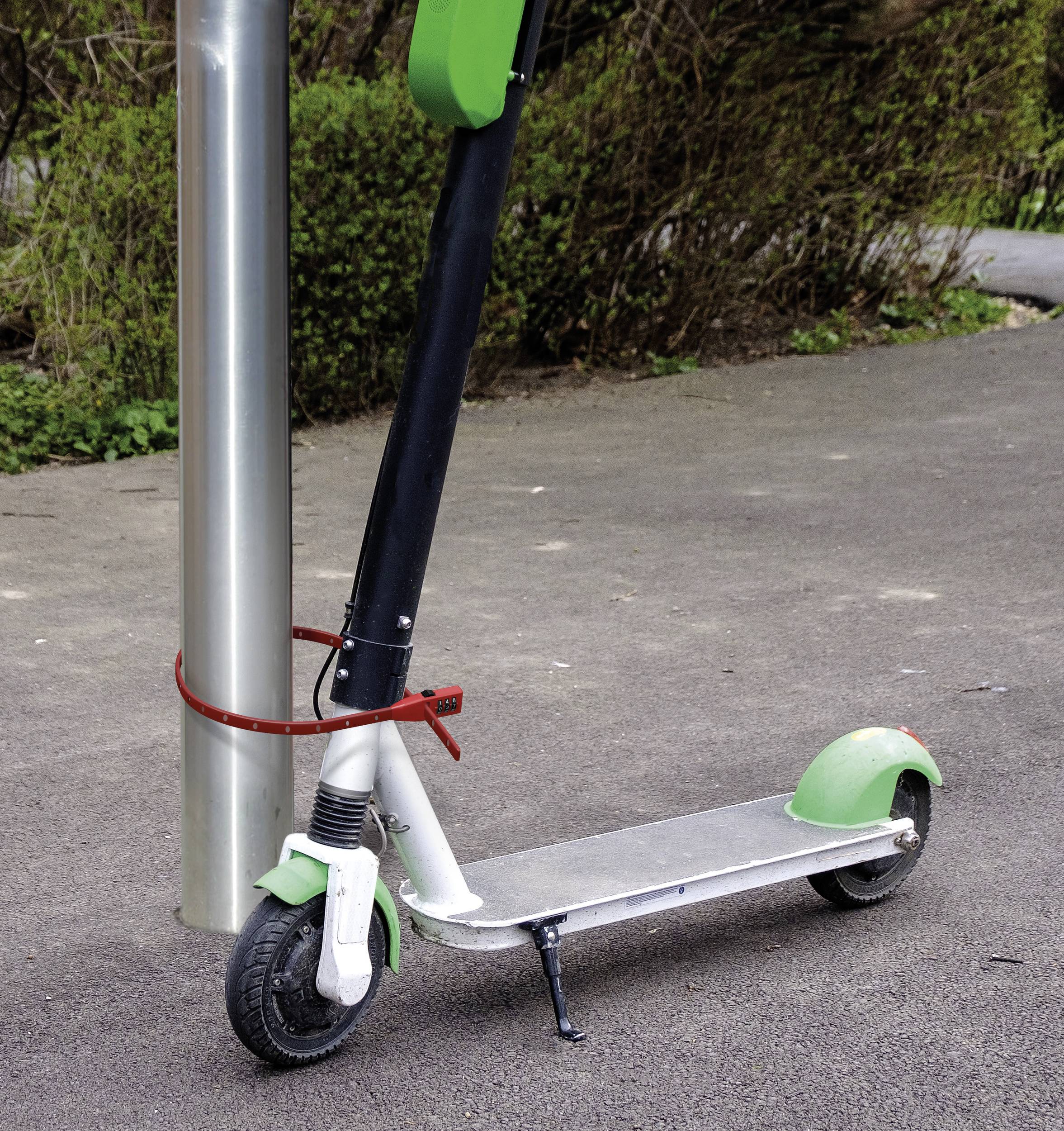 Elektryczna hulajnoga jest przymocowana czerwonym linką zabezpieczającą do metalowego słupa. Stoi na wybrukowanym chodniku.