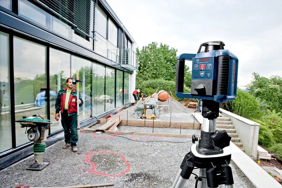 Robotnik budowlany używa urządzenia pomiarowego laserowego na placu budowy. W tle widoczne są szyby oraz różne narzędzia budowlane.