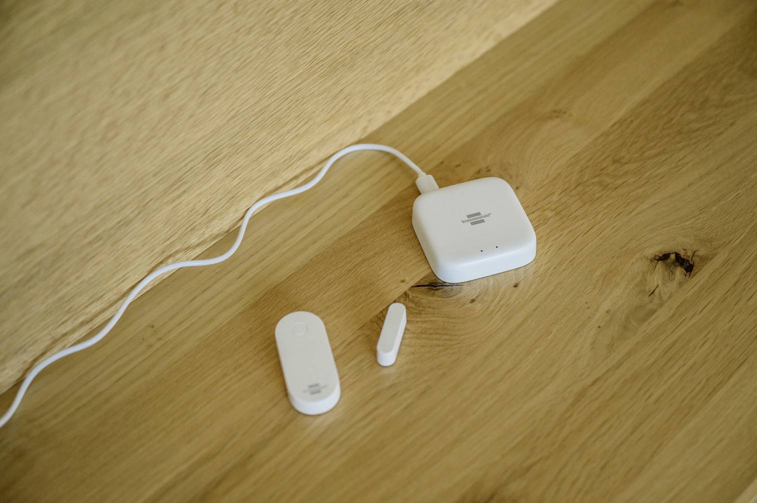 Inteligentny zestaw czujników do drzwi leży na drewnianym stole, prezentując główny czujnik, kontakt magnetyczny oraz centrum sterujące z podłączonym przewodem.