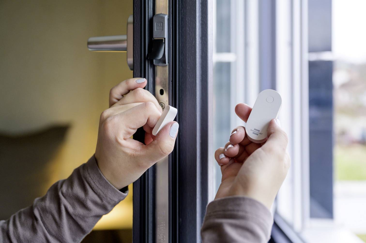 Osoba trzyma dwa białe czujniki przy otwartej framudze okiennej.