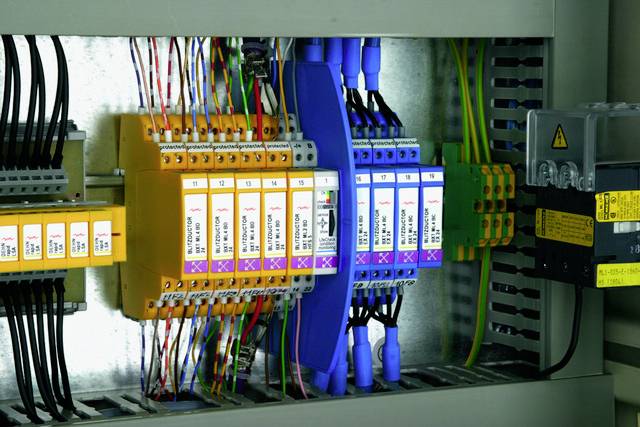 Skrzynka elektryczna z kolorowymi przewodami i bezpiecznikami w różnych kolorach. Po prawej stronie znajduje się żółty znak ostrzegawczy.