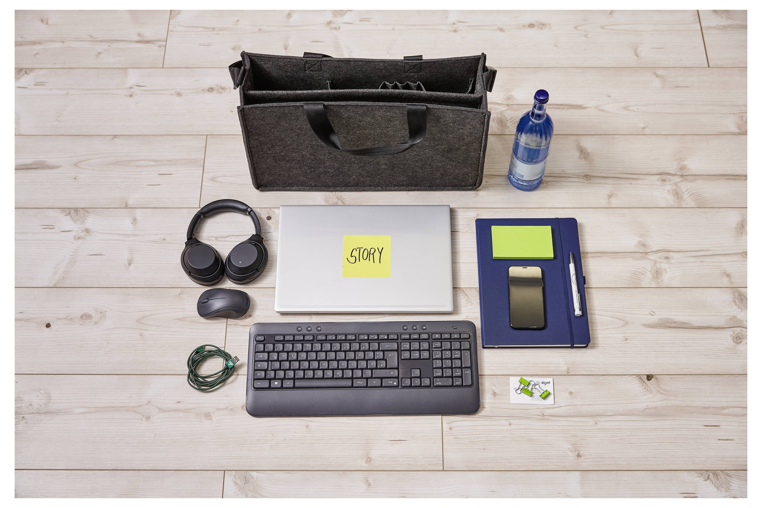 Flat lay pracowni na drewnianej podłodze: laptop oznaczony „STORY" z klawiaturą, słuchawkami, myszką, notatnikiem z długopisem, butelką wody, torbą i kablami.