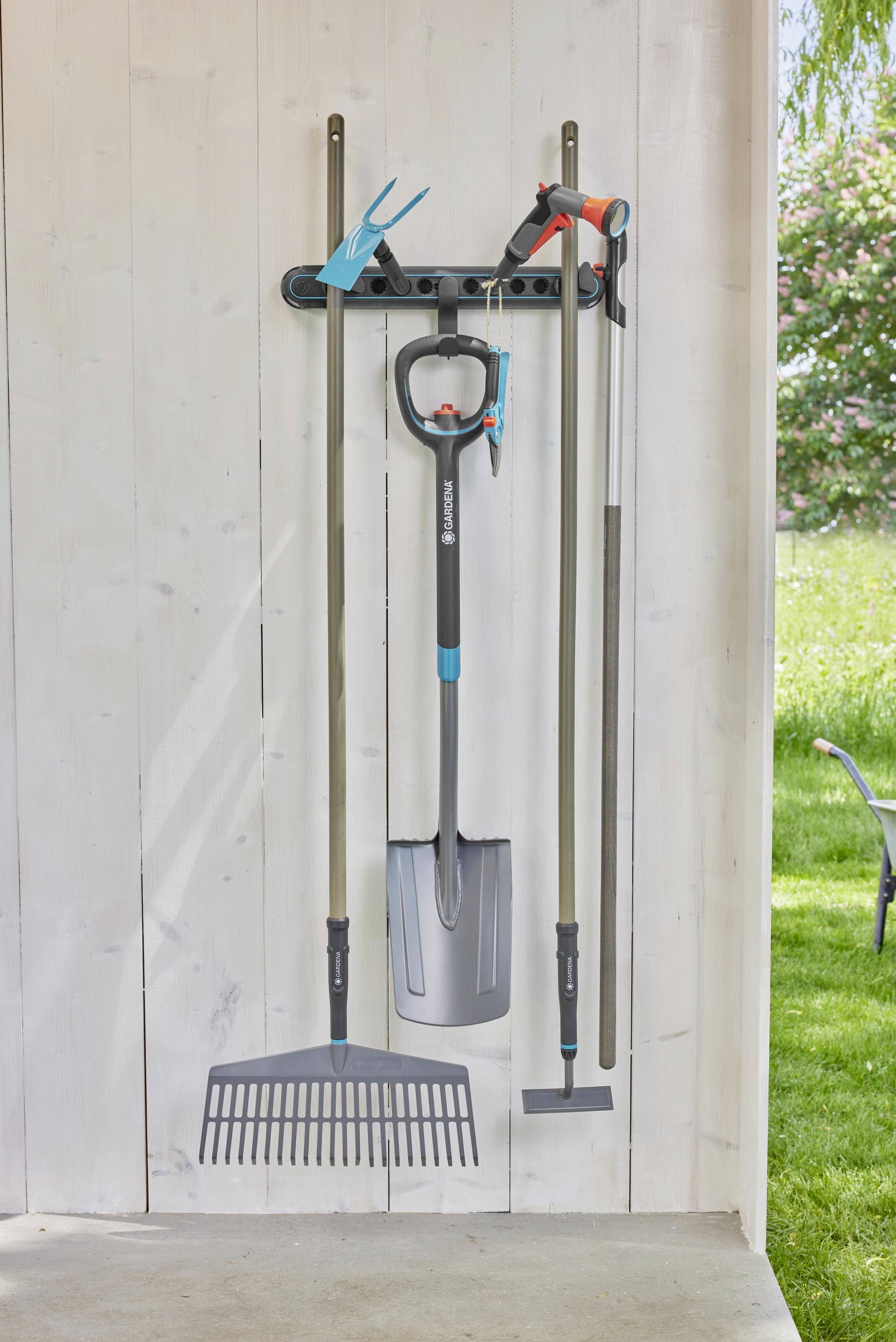 Narzędzia ogrodnicze, w tym grabie, łopata i szpadel, wiszą na białej drewnianej ścianie w strefie ogrodowej.