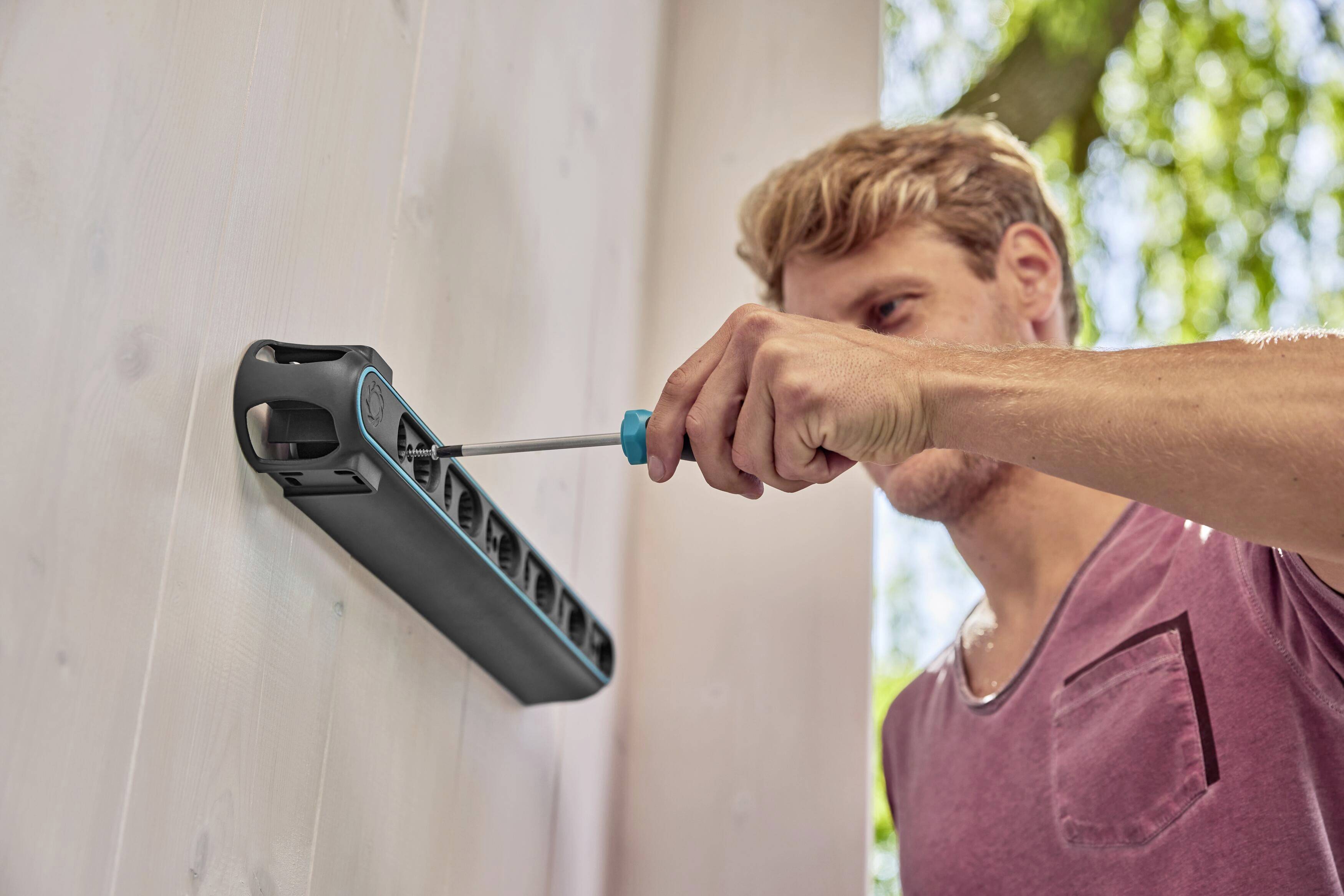 Mężczyzna montuje czarny system nawadniania Gardena na drewnianej ścianie za pomocą śrubokręta pod słonecznym niebem.