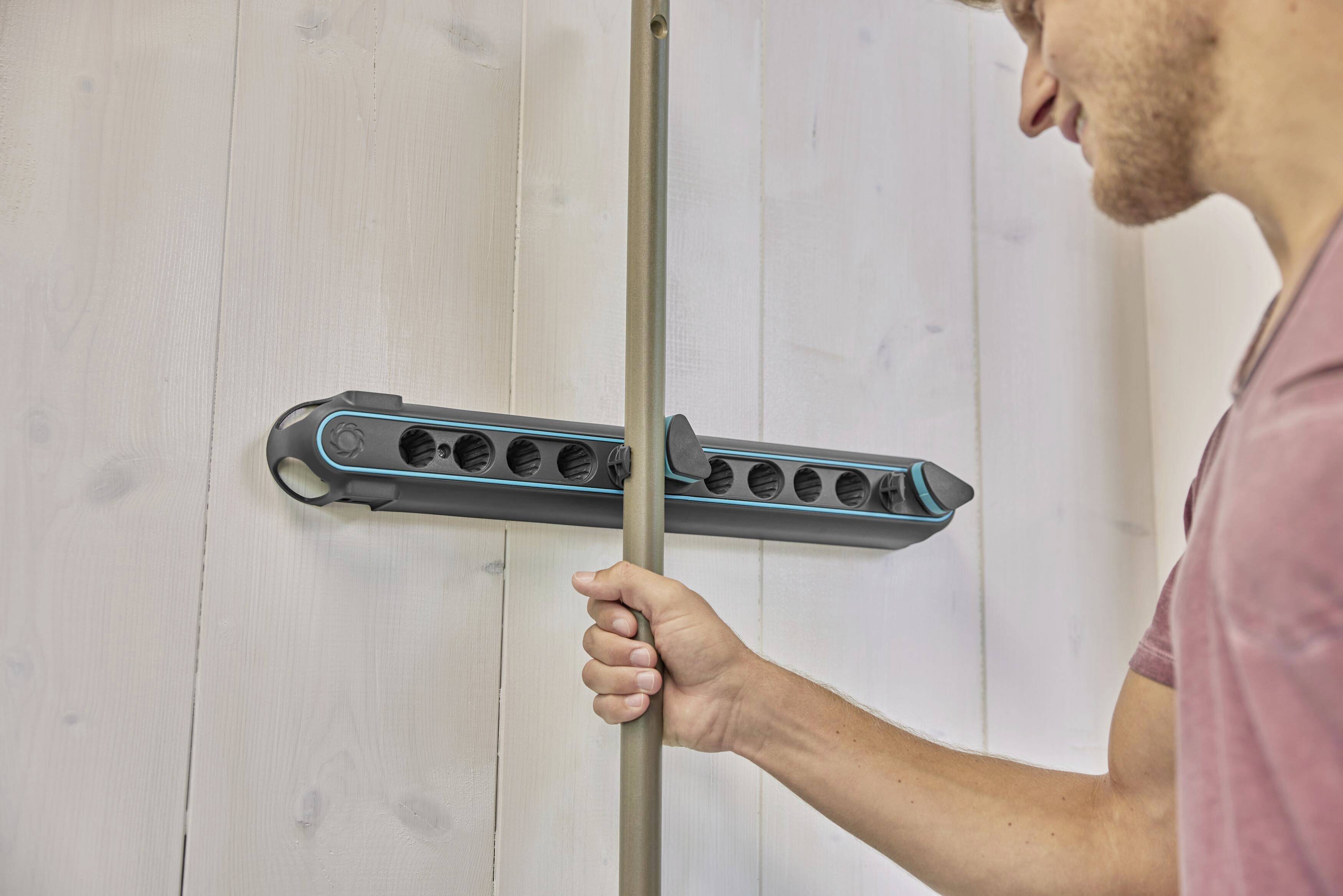 Osoba mocuje szarą rurę na wieszaku ściennym z wieloma uchwytami, prawdopodobnie w celu przechowywania sprzętów gospodarstwa domowego.
