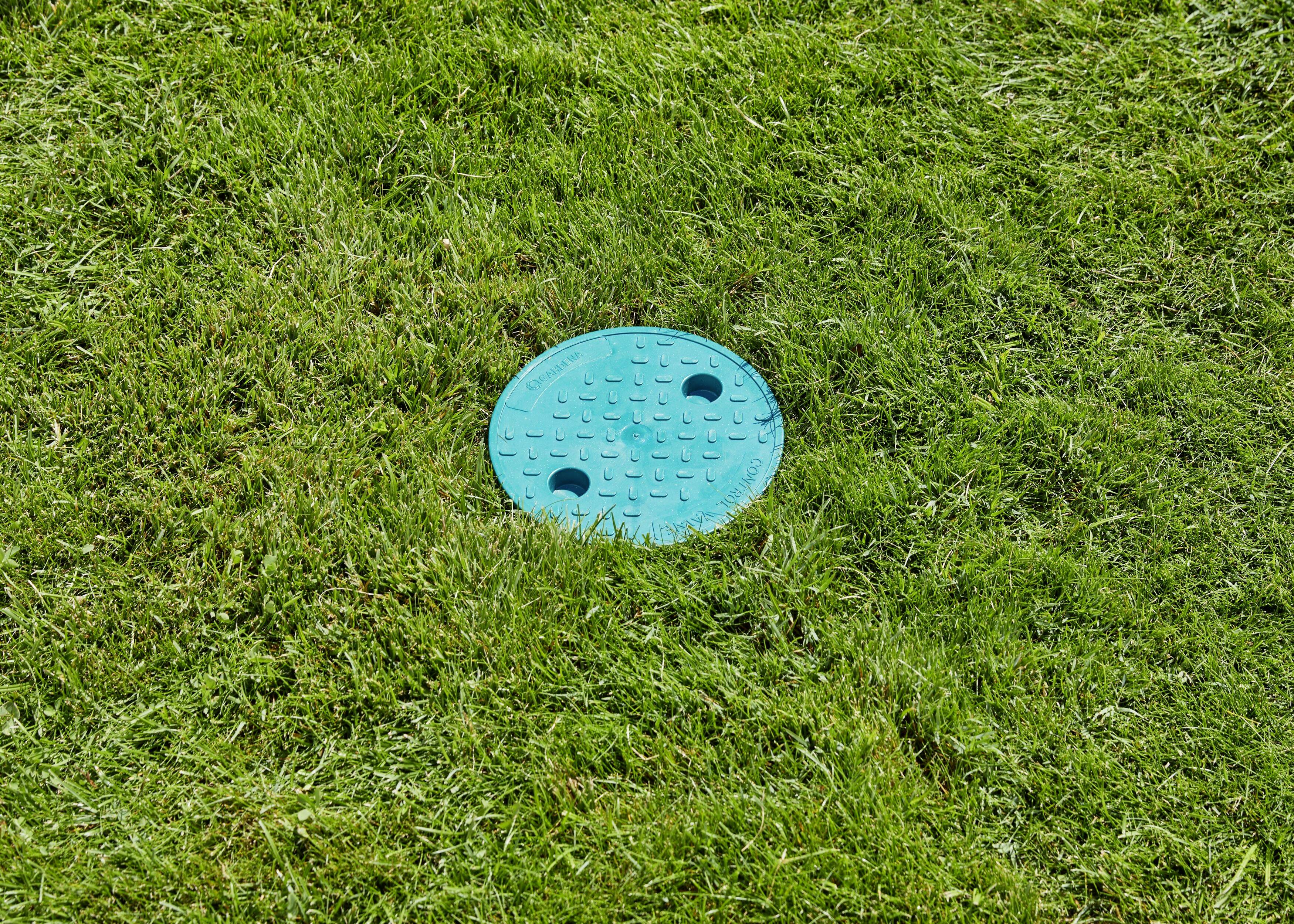 Niebieski właz kanalizacyjny na zielonym trawniku. Pokrywa posiada dwa otwory i jest otoczona przyciętą trawą.