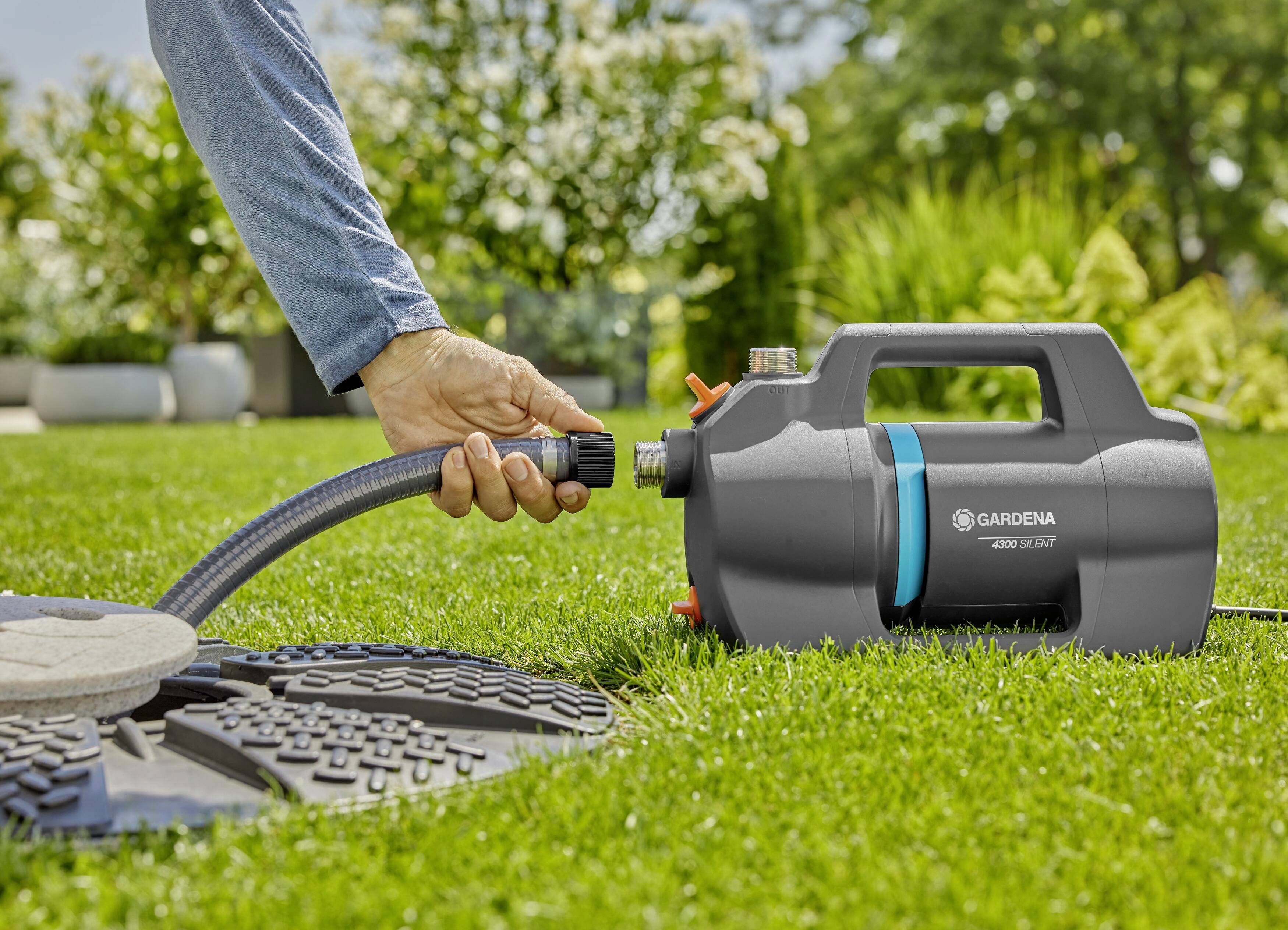 Osoba podłącza wąż ogrodowy do pompy ogrodowej GARDENA na zielonej trawnicy; w tle widoczne są drzewa i krzewy.