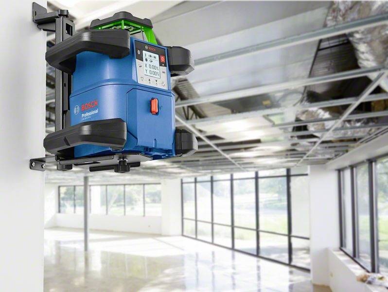 Niebieski laserowy niwelator budowlany jest zamontowany na metalowej konstrukcji w niedokończonym pomieszczeniu. Przez okna wpada światło dzienne.