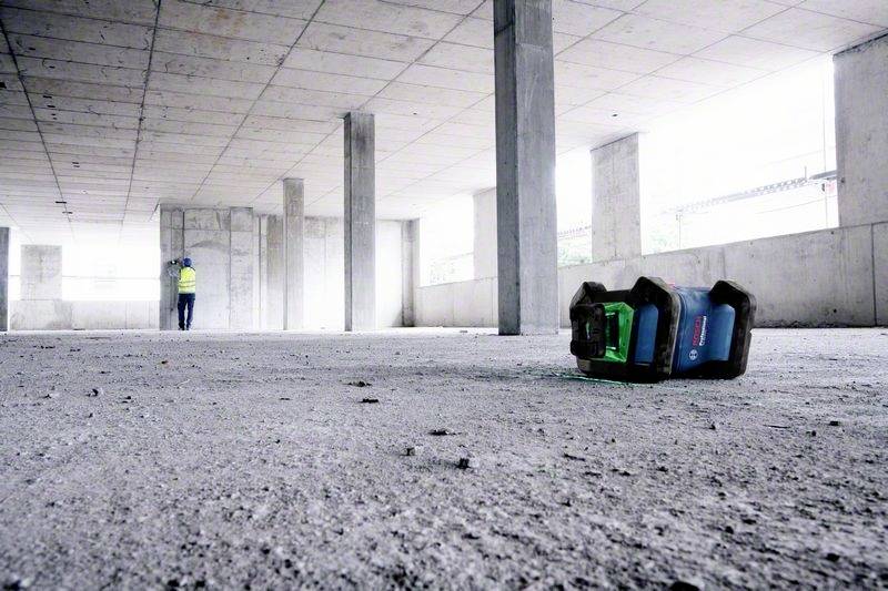 Robotnik budowlany w kamizelce odblaskowej stoi w niedokończonym budynku betonowym. Laserowy przyrząd do niwelacji leży na pierwszym planie na podłodze.