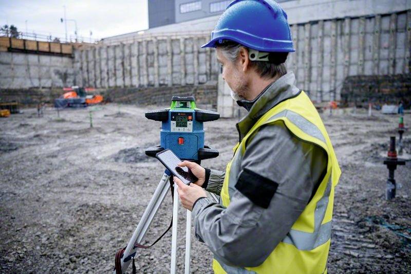 Robotnik budowlany w kasku i kamizelce odblaskowej obsługuje urządzenie pomiarowe na placu budowy. W tle trwają prace budowlane.