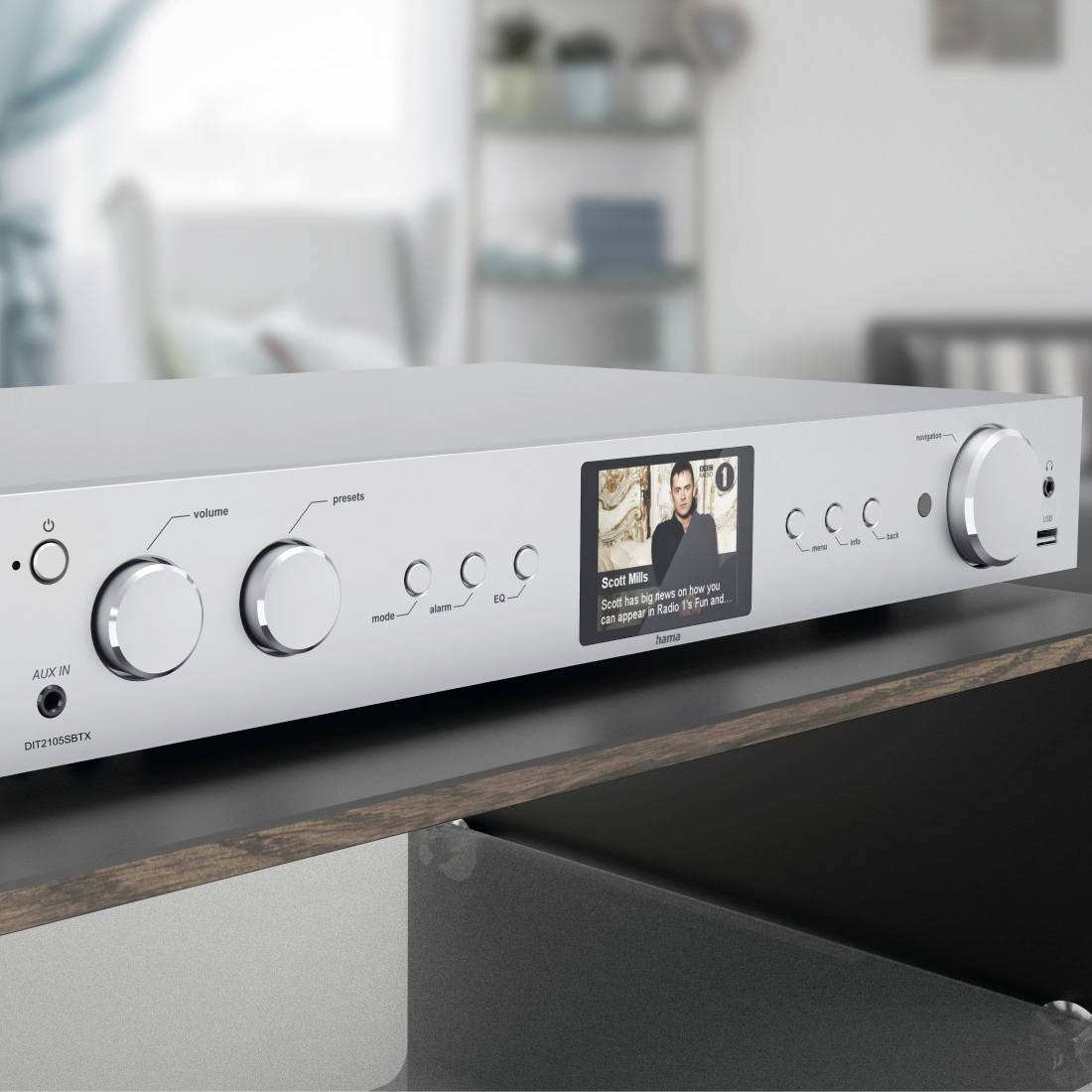 Srebrny wzmacniacz z wieloma regulatorami i małym wyświetlaczem pokazującym tytuł utworu muzycznego. W tle widoczny jest salon.