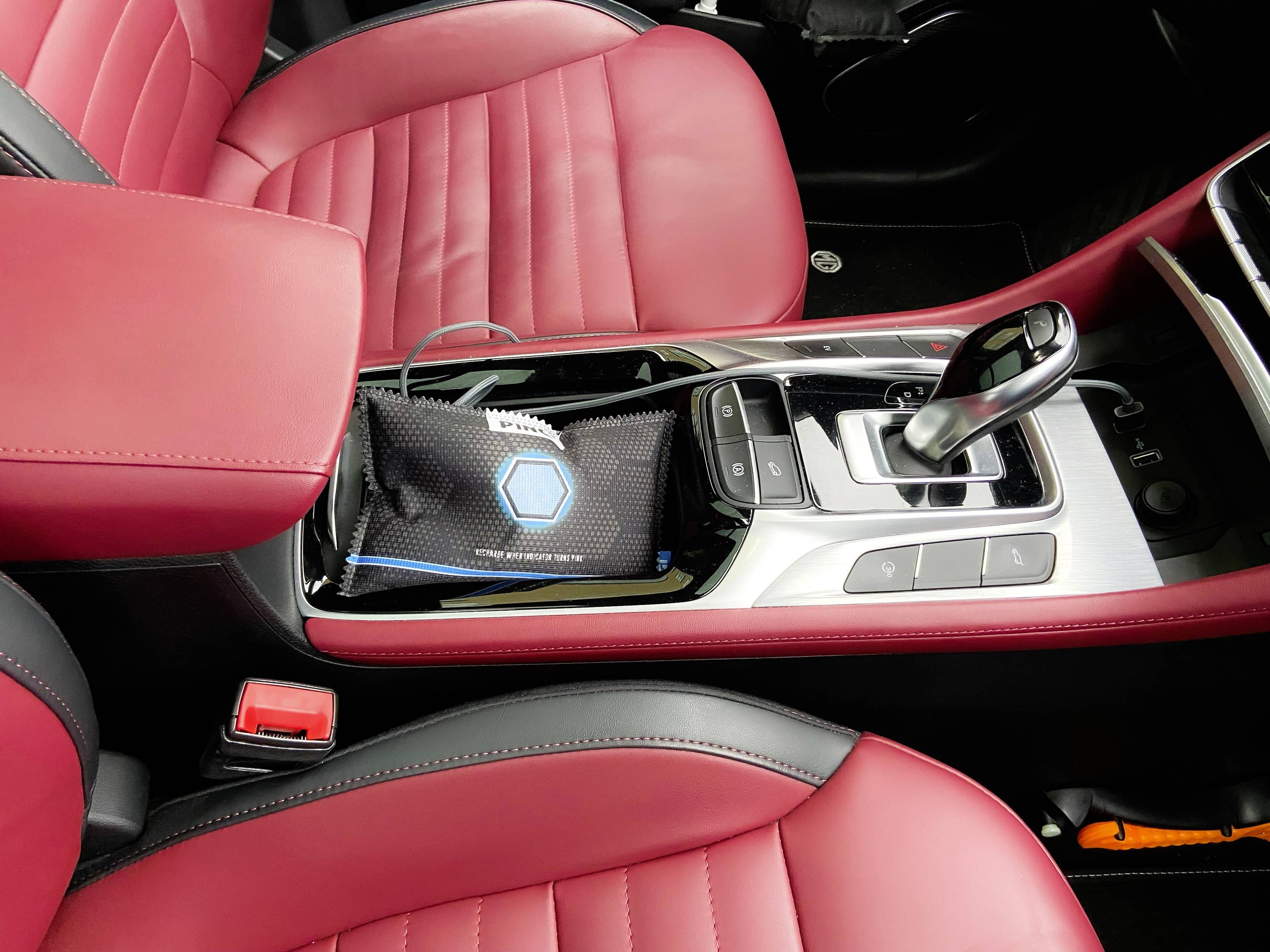 Czerwony wnętrze siedzeń samochodu z czarną konsolą środkową, zawierające czarną torbę. Dźwignia zmiany biegów widoczna między fotelami przednimi.