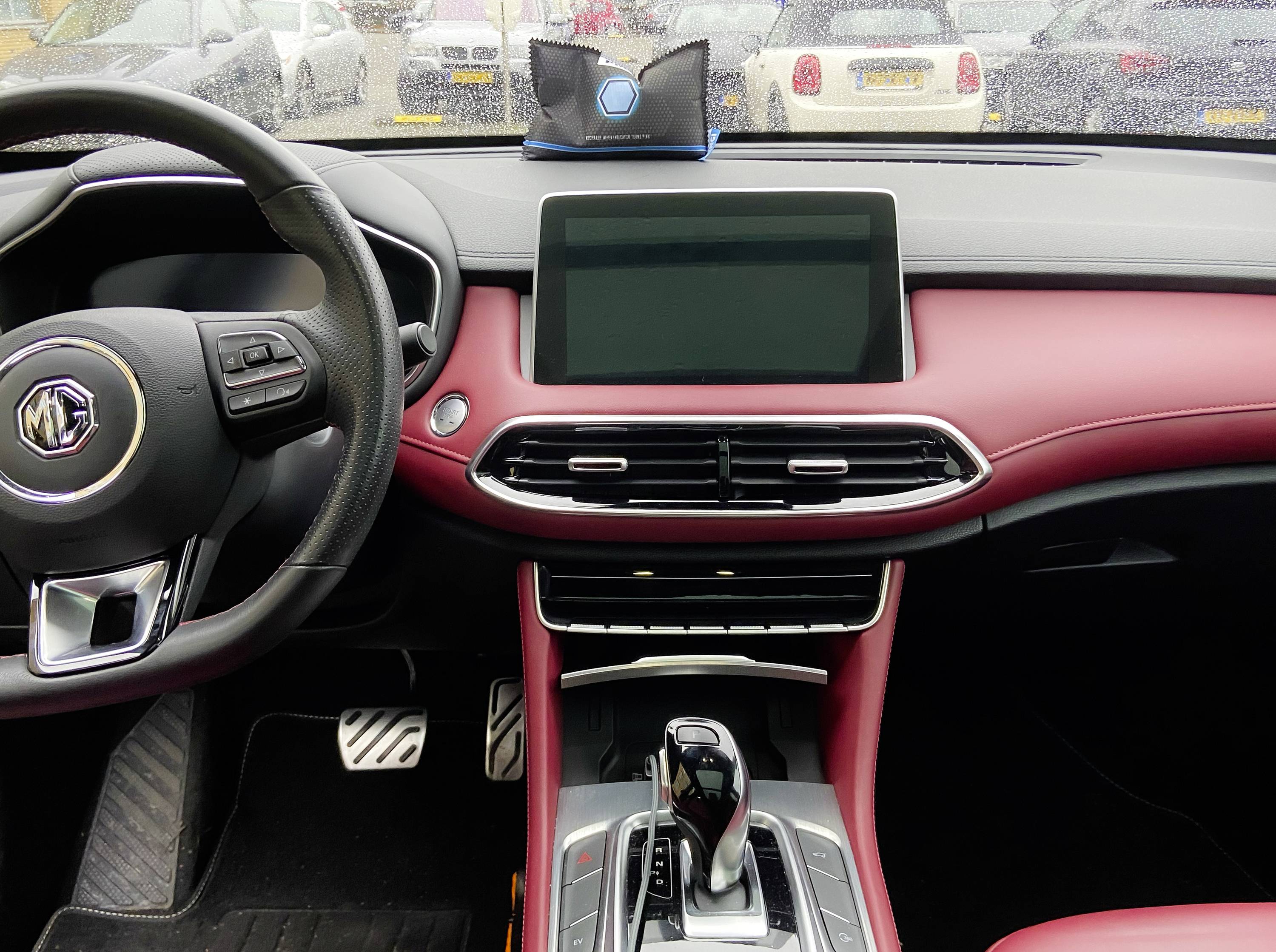 Wnętrze nowoczesnego samochodu: kierownica po lewej stronie, wyświetlacz pośrodku, deska rozdzielcza w kolorach czerwonym i czarnym. W tle zaparkowane są samochody.