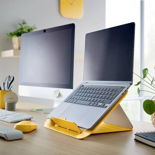 Dwa laptopy stoją na biurku w biurze, z żółtymi akcesoriami takimi jak mysz i podstawka pod laptopa. W tle widać okna.