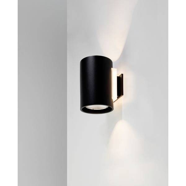 Czarna, cylindryczna lampa ścienna na białej ścianie, oświetlająca przestrzeń w górę i w dół.