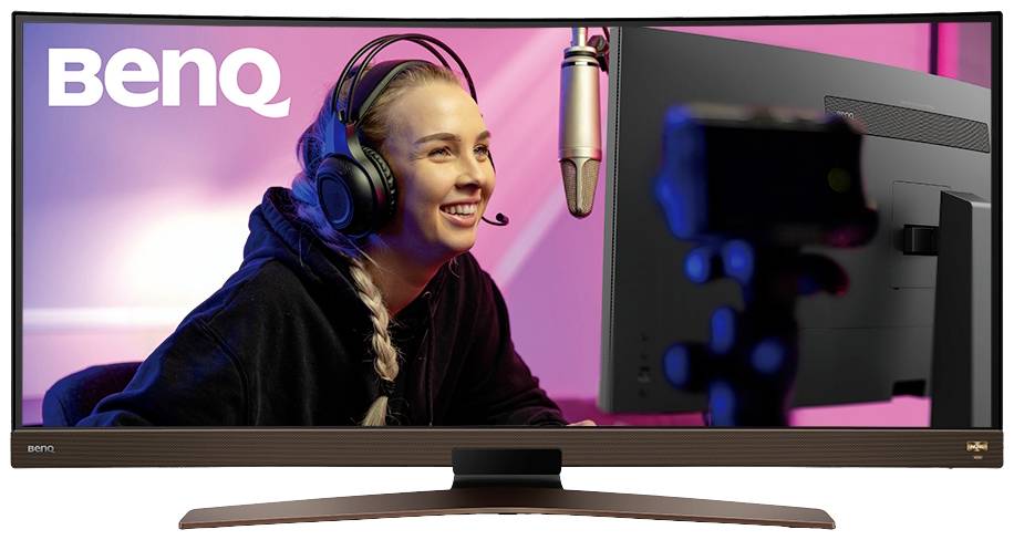 Osoba w słuchawkach siedzi uśmiechnięta przed mikrofonem i ekranem komputera. W tle widać napis 'BenQ'.