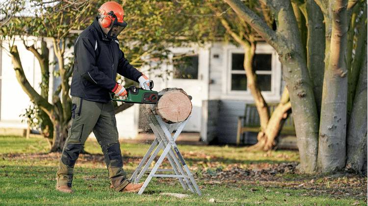 Osoba w odzieży ochronnej używa piły mechanicznej do cięcia dużego pnia drzewa na łące, z małym domkiem w tle.