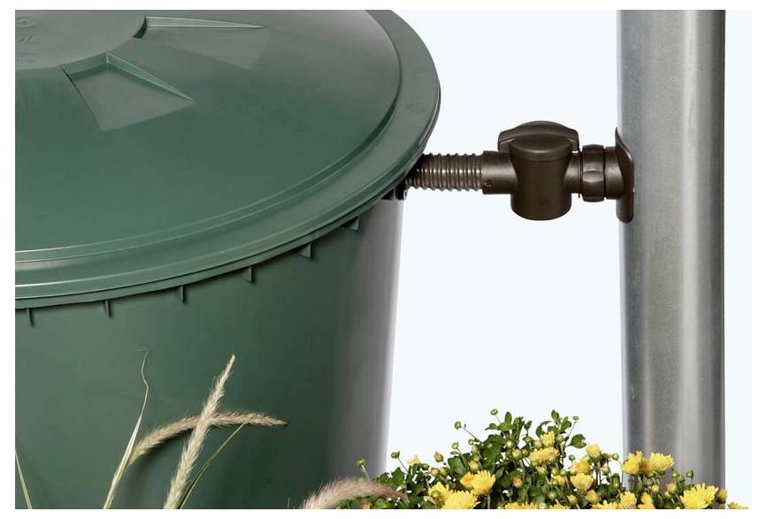 Zielony kosz na śmieci zamocowany na metalowym słupie, z kwiatami u dołu na tle jednolitego, jasnego tła.