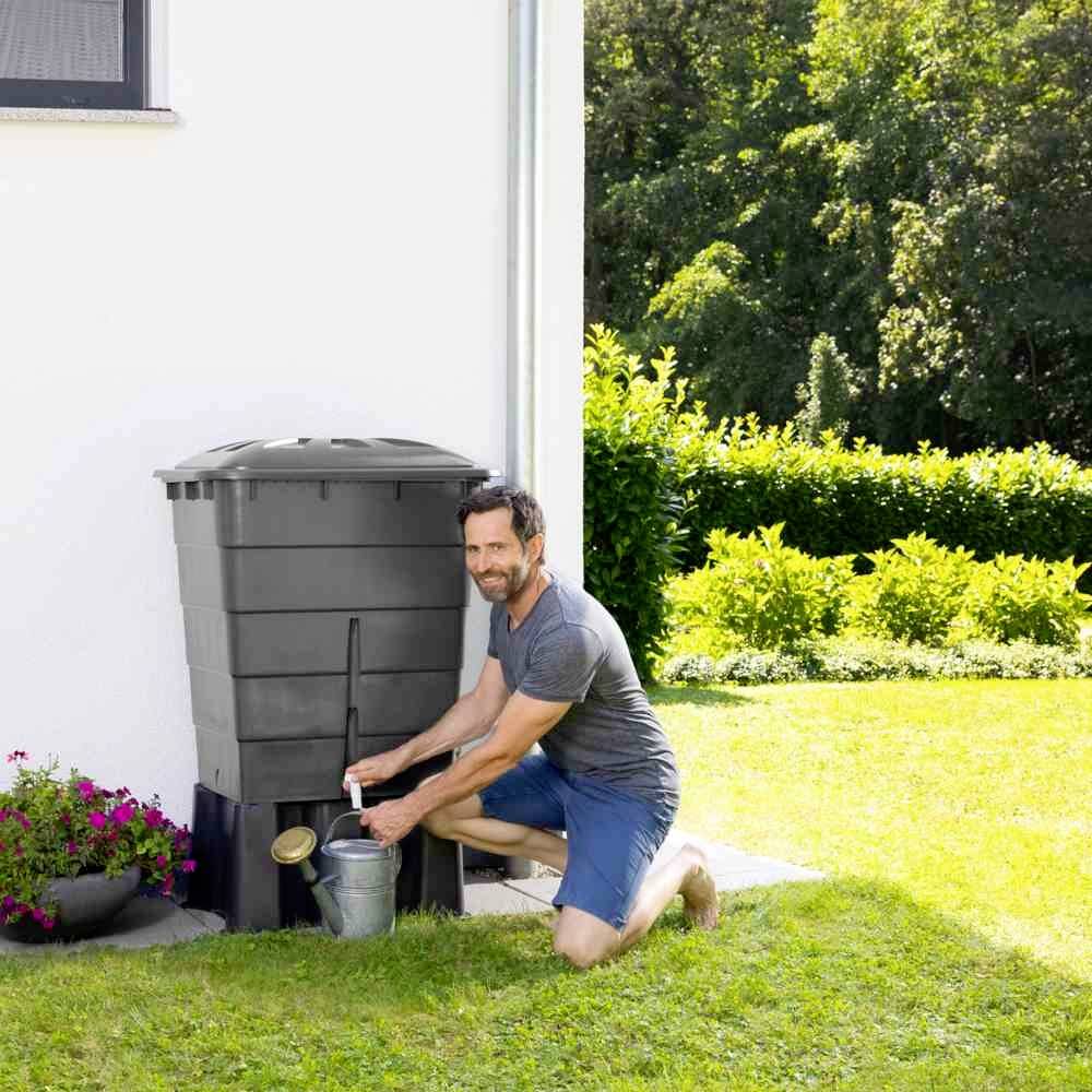 Mężczyzna napełnia konewkę wodą z beczki na deszczówkę w słoneczny dzień w ogrodzie. W tle krzewy i trawnik.
