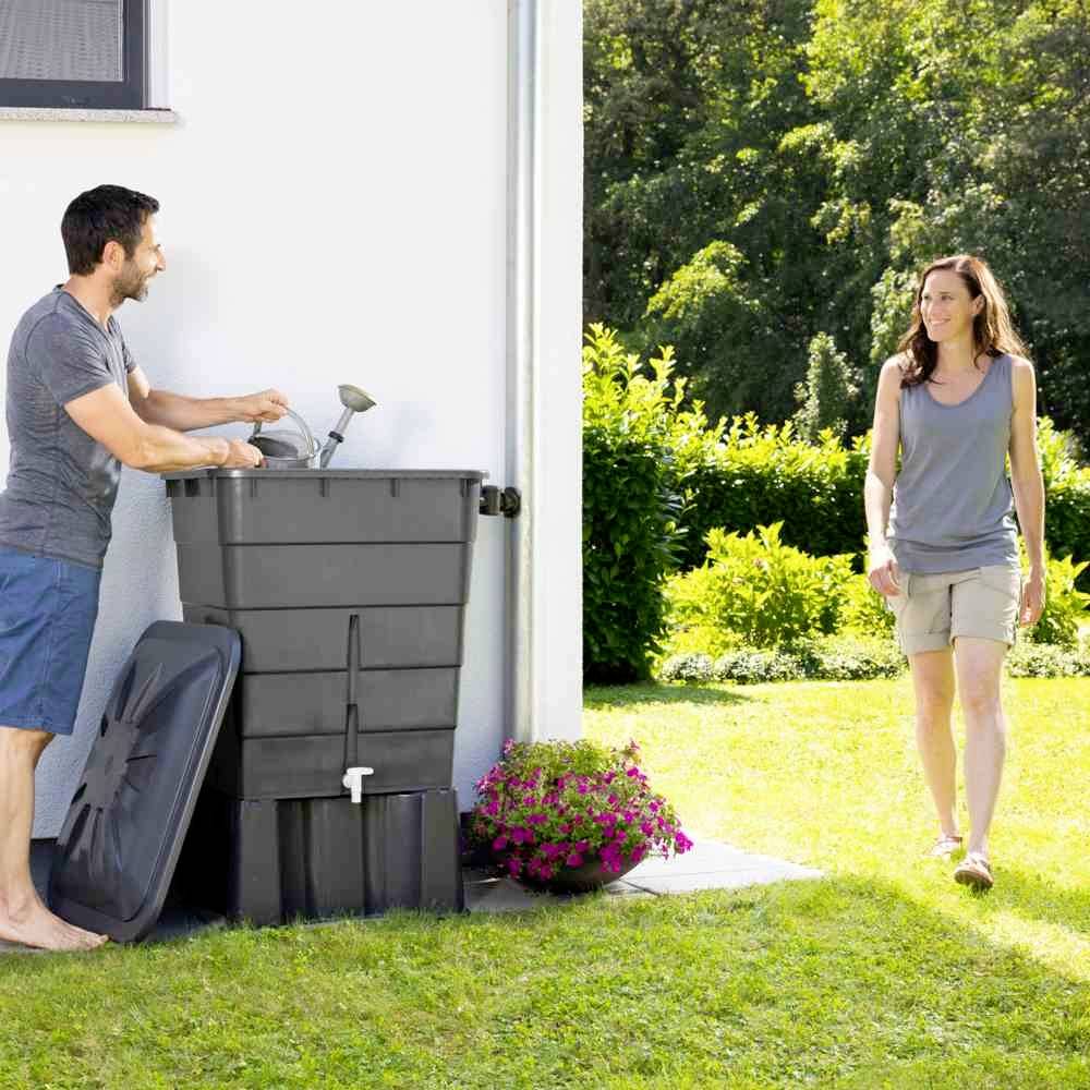 Mężczyzna stoi obok dużej beczki na deszczówkę i śmieje się, podczas gdy kobieta zbliża się do niego na łące.