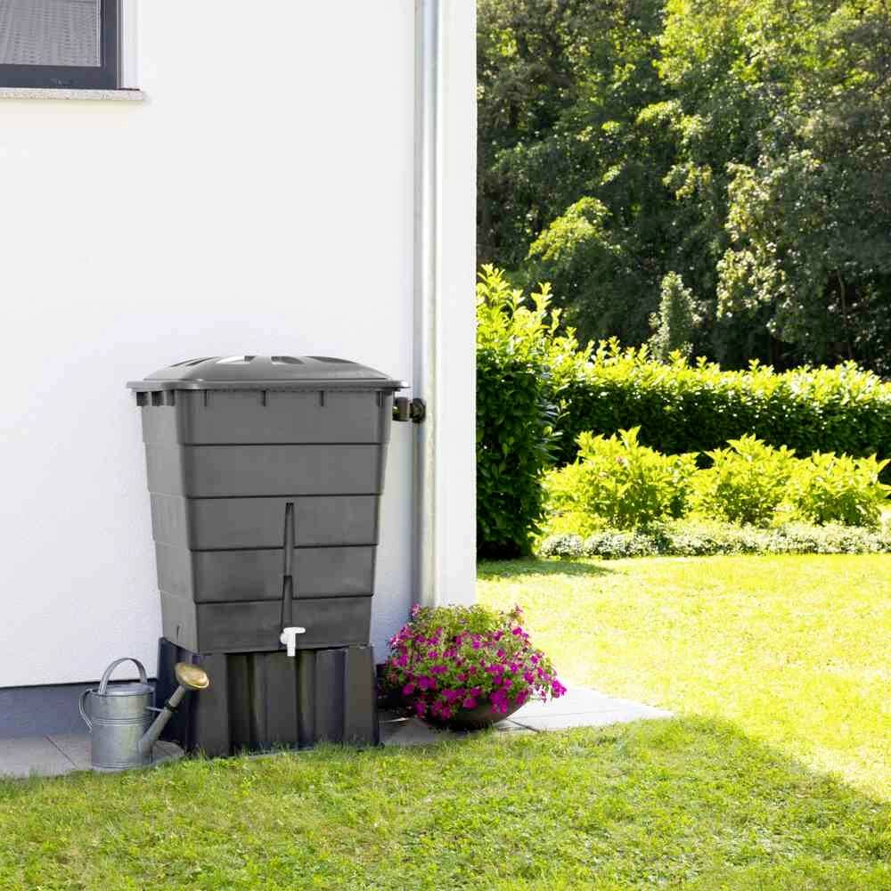 Zbiornik na deszczówkę z kranem stoi obok domu, otoczony ogrodem z zieloną trawą i kwitnącymi roślinami.