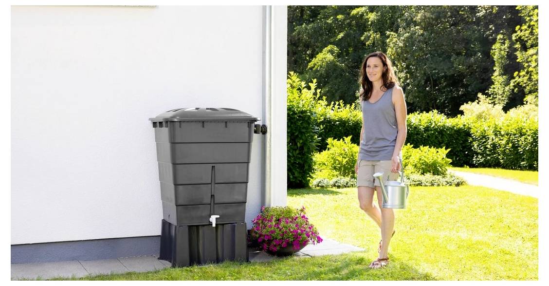 Kobieta niosąca konewkę przechodzi obok beczki na deszczówkę stojącej przy domu, z ogrodem i kwiatami w tle.