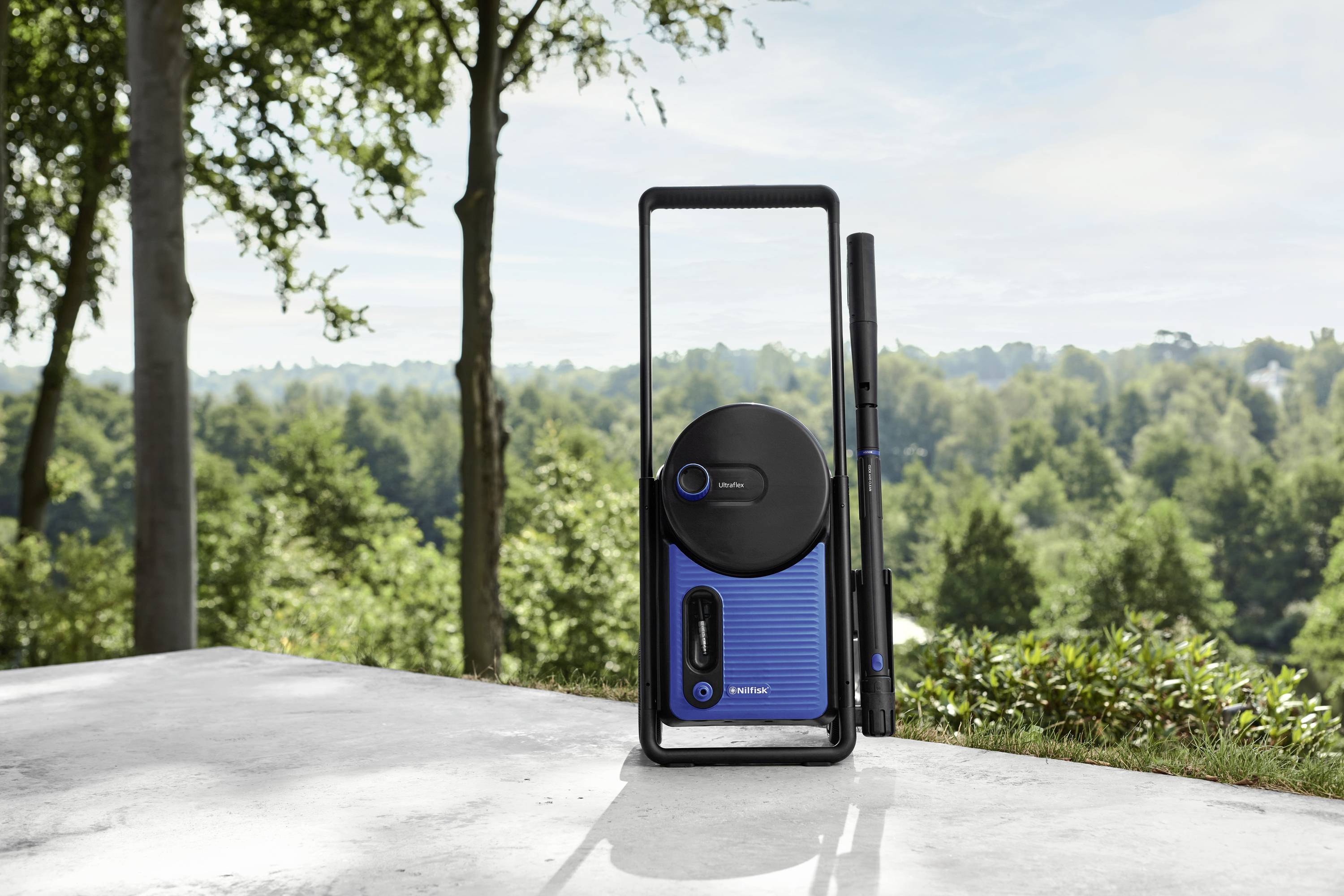 Przenośny system głośnikowy w niebieskiej obudowie stoi na tarasie z widokiem na zalesiony wzgórze w tle.