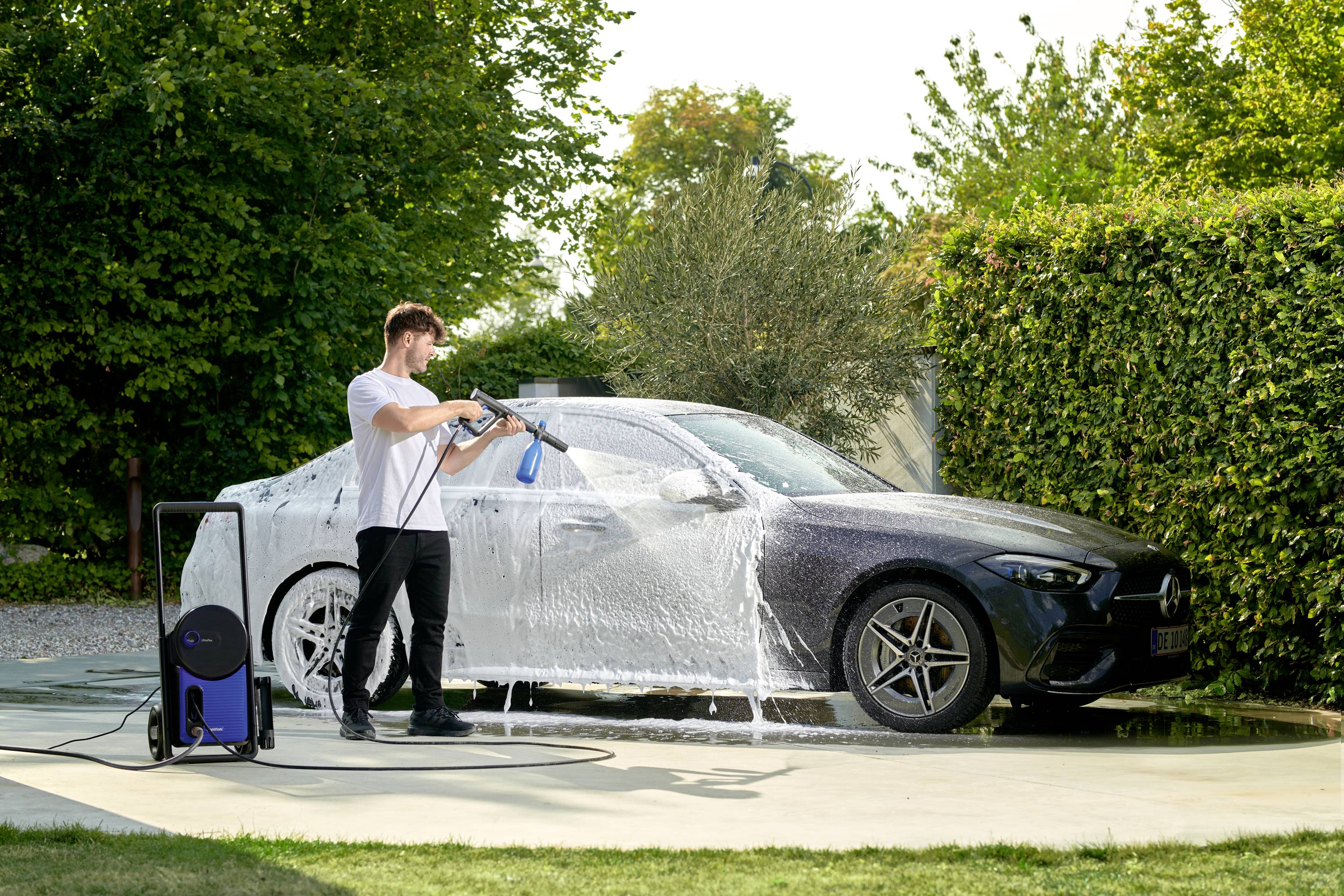 Mężczyzna myje samochód myjką ciśnieniową w ogrodzie. Samochód jest pokryty pianą mydlaną. W tle widoczne są krzewy.