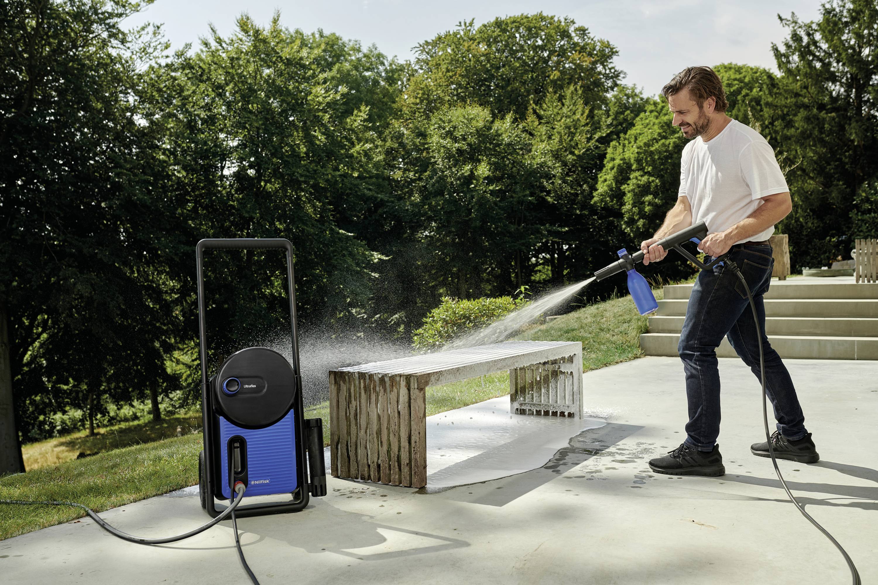 Mężczyzna czyści zewnętrzną ławkę myjką ciśnieniową na tarasie, w tle otoczony drzewami.