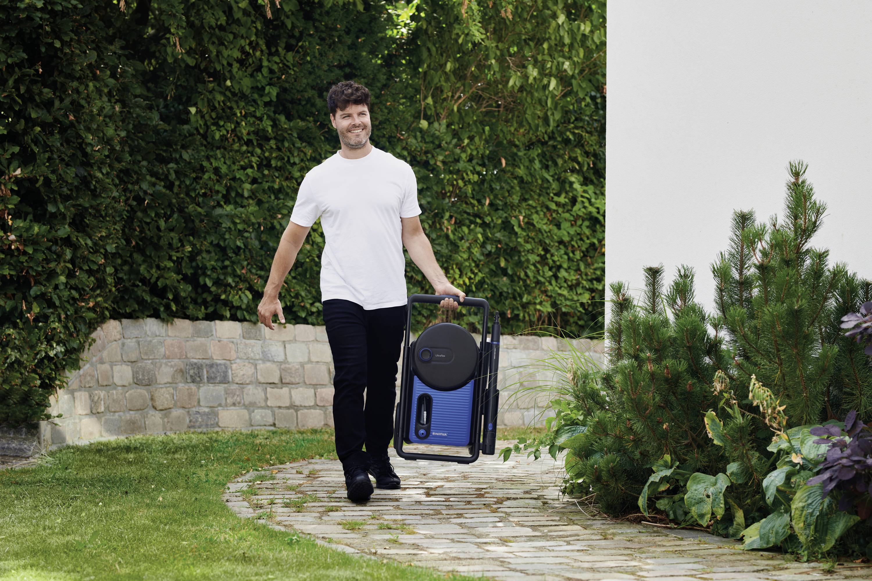 Mężczyzna uśmiechając się idzie po wybrukowanej ścieżce w ogrodzie. Niesie przenośny, niebieski, prostokątny przedmiot. W tle widać gęsty zielony żywopłot.