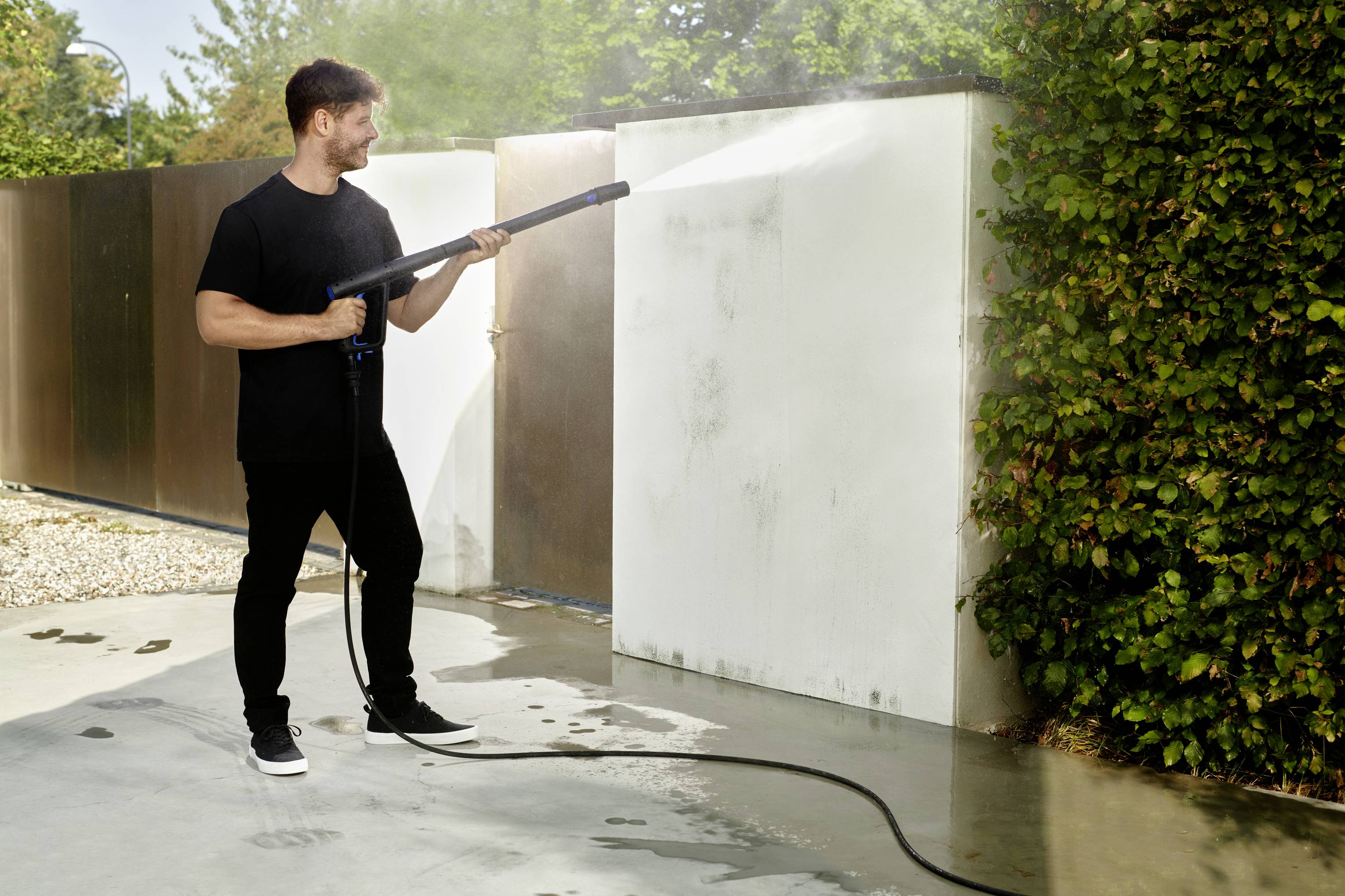 Mężczyzna czyści ścianę myjką ciśnieniową na zewnątrz. Neutralna kolorystyka, zielone rośliny w tle.