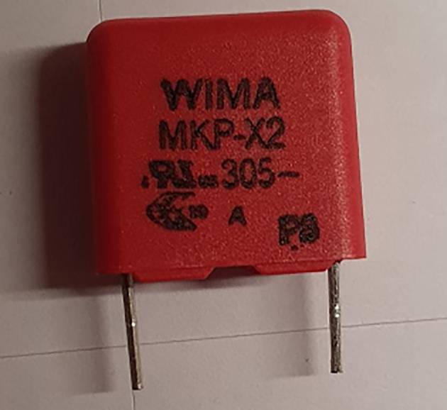 Czerwony kondensator WIMA MKP-X2 z dwoma metalowymi pinami, leżący na białej powierzchni, pokazujący informacje techniczne na swojej przedniej stronie.
