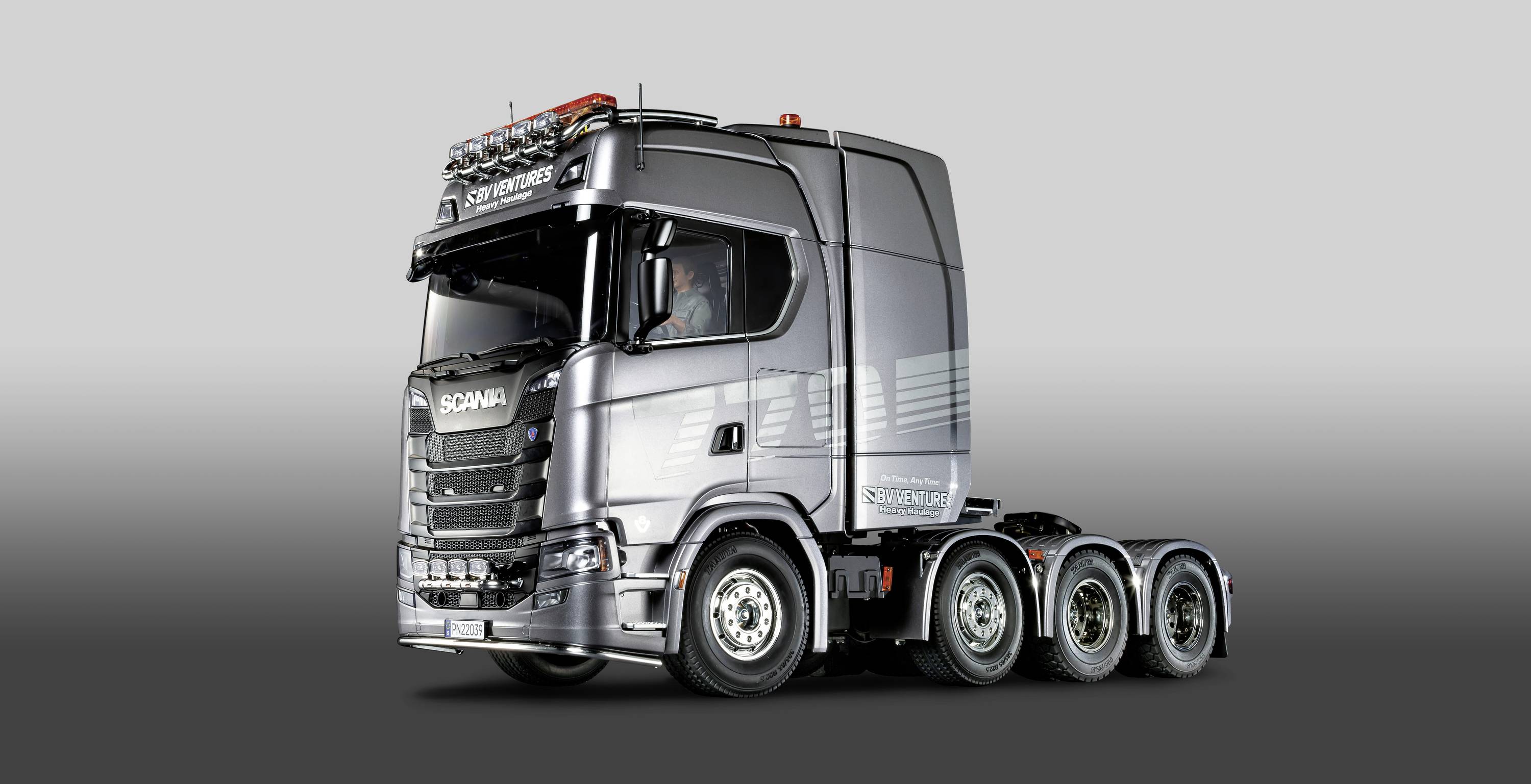 Srebrny samochód ciężarowy marki Scania z czterema osiami, sfotografowany z boku, na szarym tle.