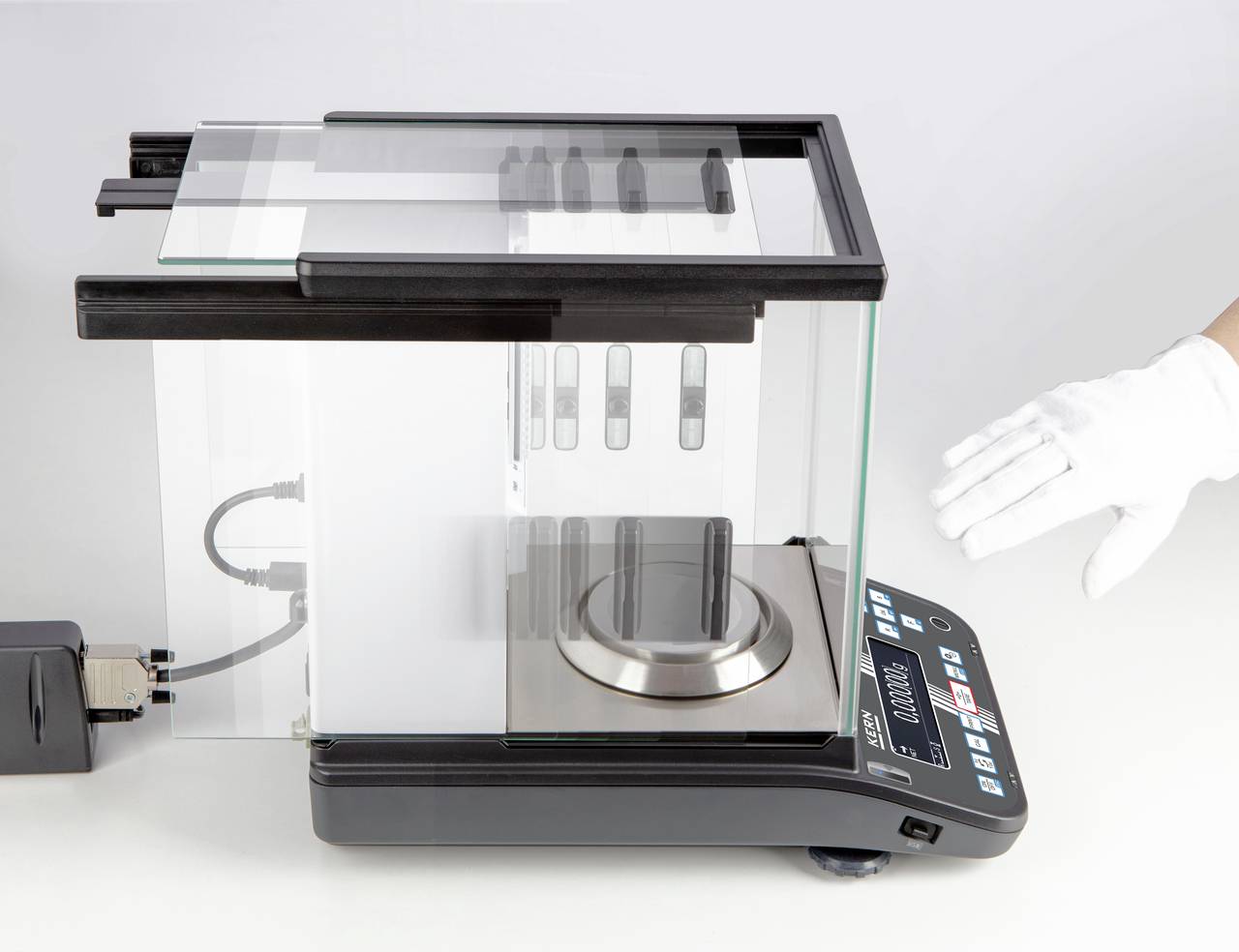 Elektroniczna waga precyzyjna w szklanej obudowie, z podłączonym przewodem, a obok osoba w rękawiczce wskazująca na nią palcem wskazującym.