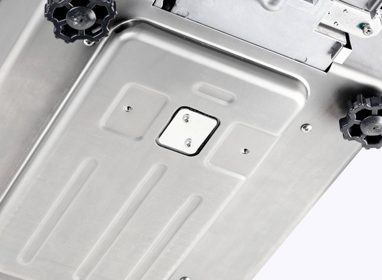 Spód urządzenia metalowego z widocznymi elementami mocującymi i dwoma czarnymi kołami, prawdopodobnie urządzenie elektryczne.