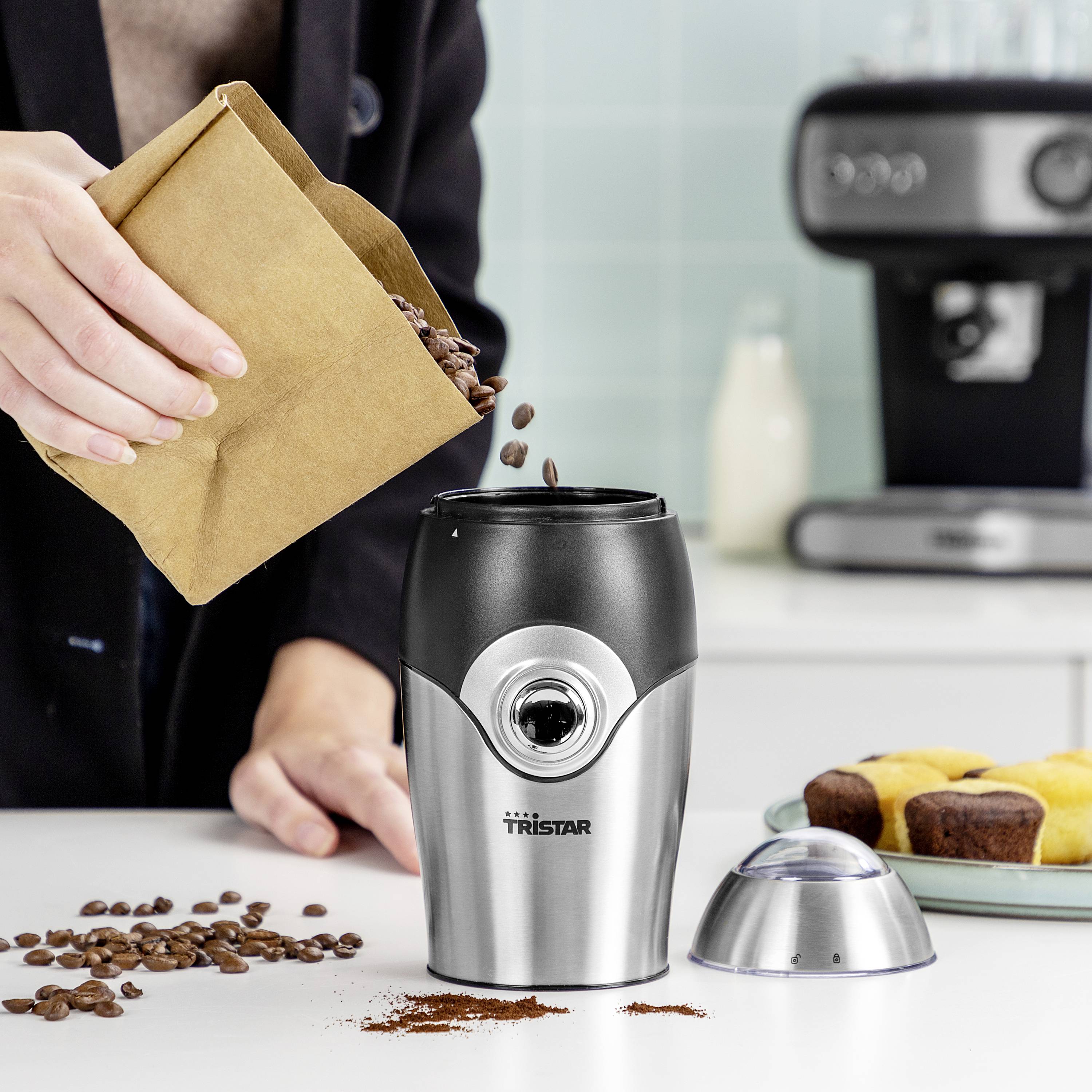 Osoba wsypuje ziarna kawy do srebrnego młynka stojącego na blacie kuchennym. Obok leżą ziarna kawy i muffinki.