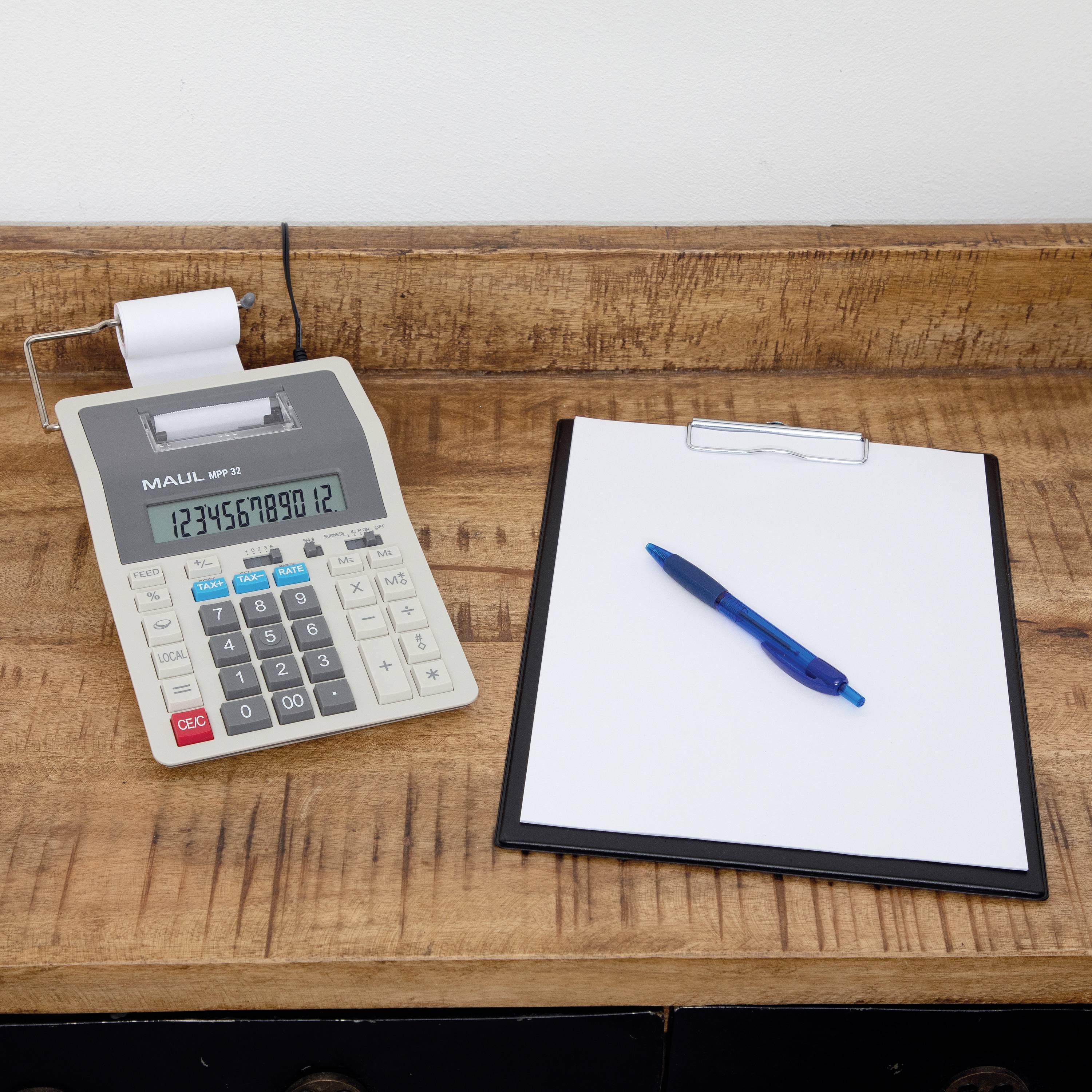 Kalkulator biurowy z rolką papieru wyświetla liczbę '789912'. Obok leży deska z klipsem i pustym arkuszem oraz niebieski długopis.