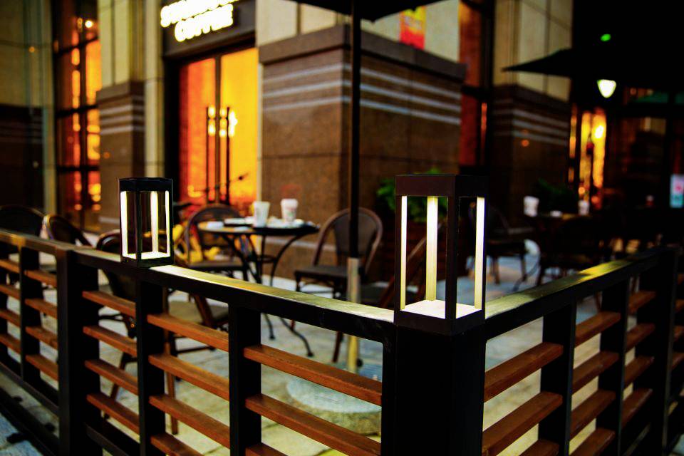 Widok z zewnątrz przytulnej kawiarni ze stolikiem i krzesłami, otoczony dekoracyjnym płotem z dwoma świecącymi latarniami.