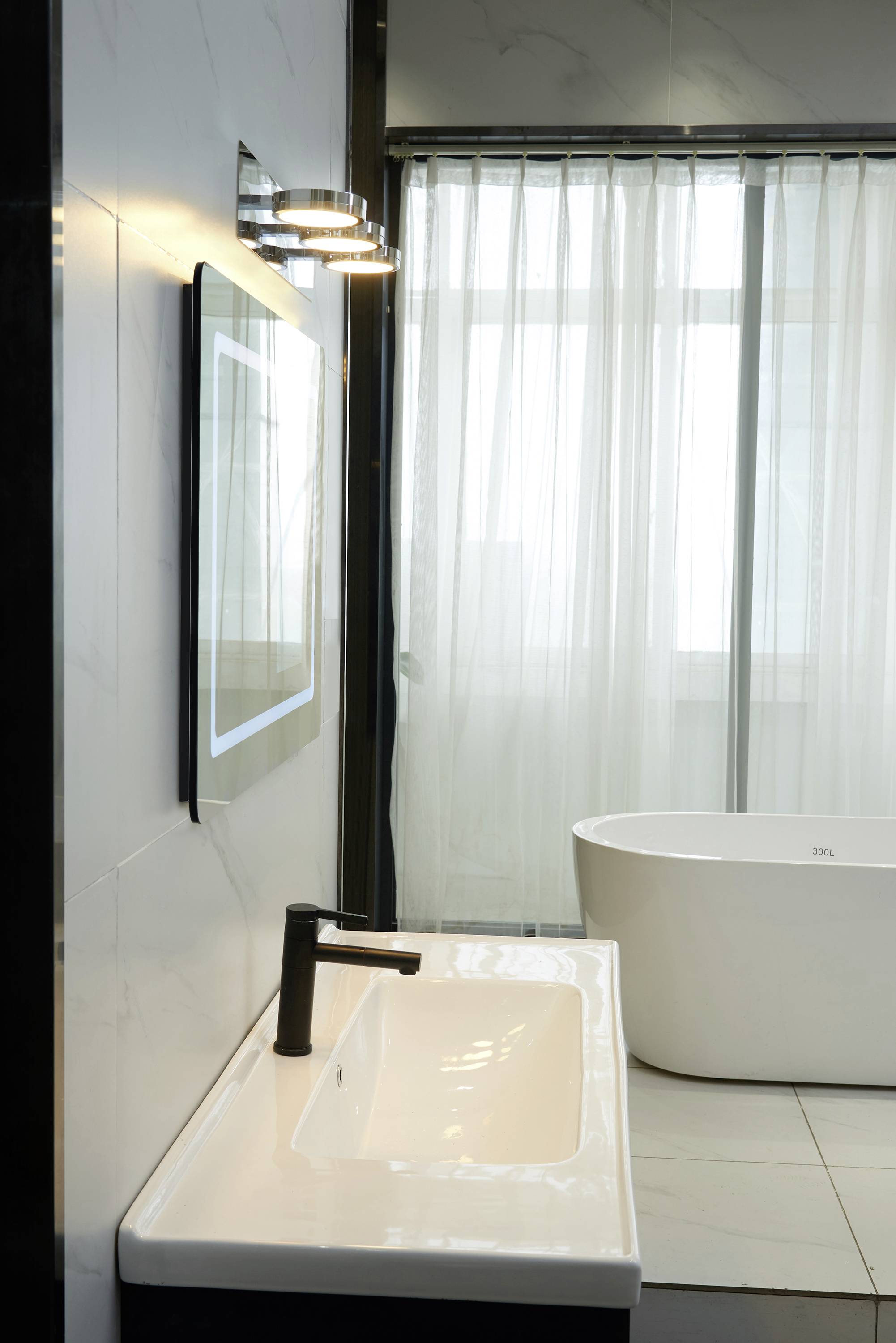 Nowoczesna łazienka z podświetlonym lustrem, umywalką z ciemną baterią oraz wolnostojącą wanną ustawioną przed oknem.