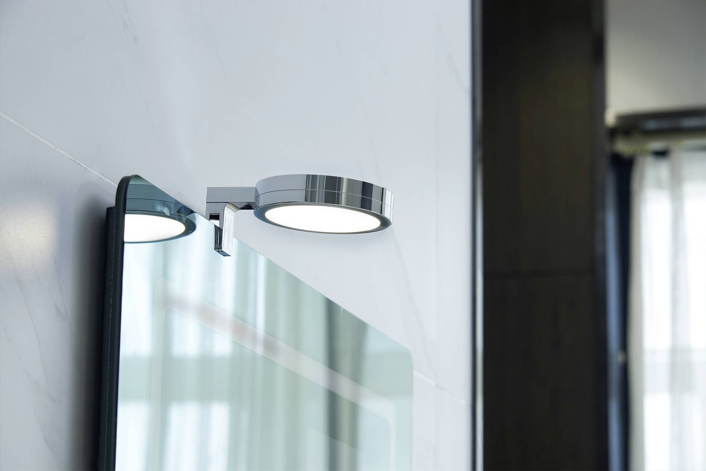 Nowoczesna okrągła lampa LED zamontowana nad lustrem na ścianie, emituje rozproszone światło, delikatnie doświetlając przestrzeń.