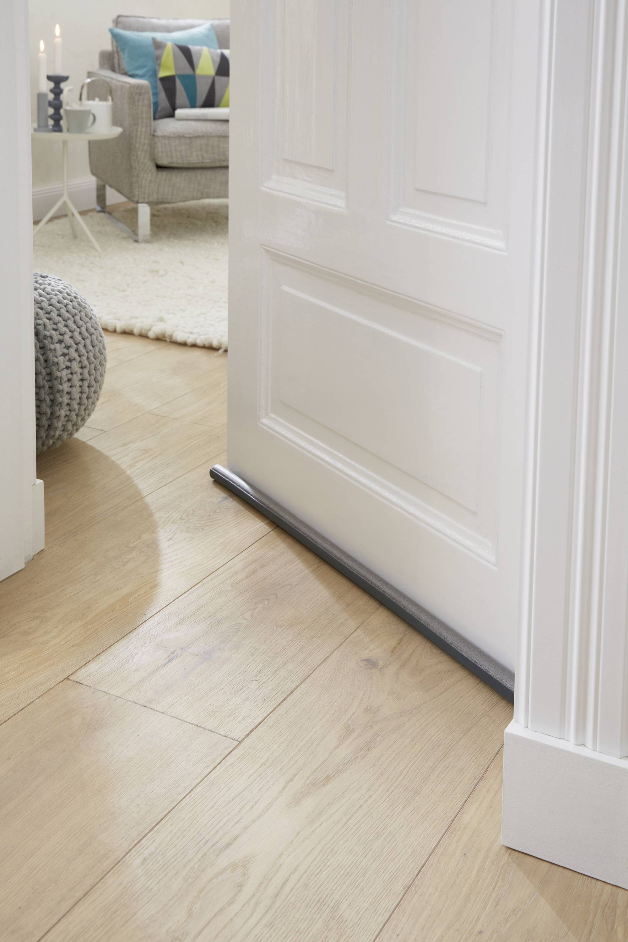 Uchylone drzwi z zatrzymywaczem ciągu powietrza przy podłodze. W tle widać przytulny salon z fotelami i dywanem.