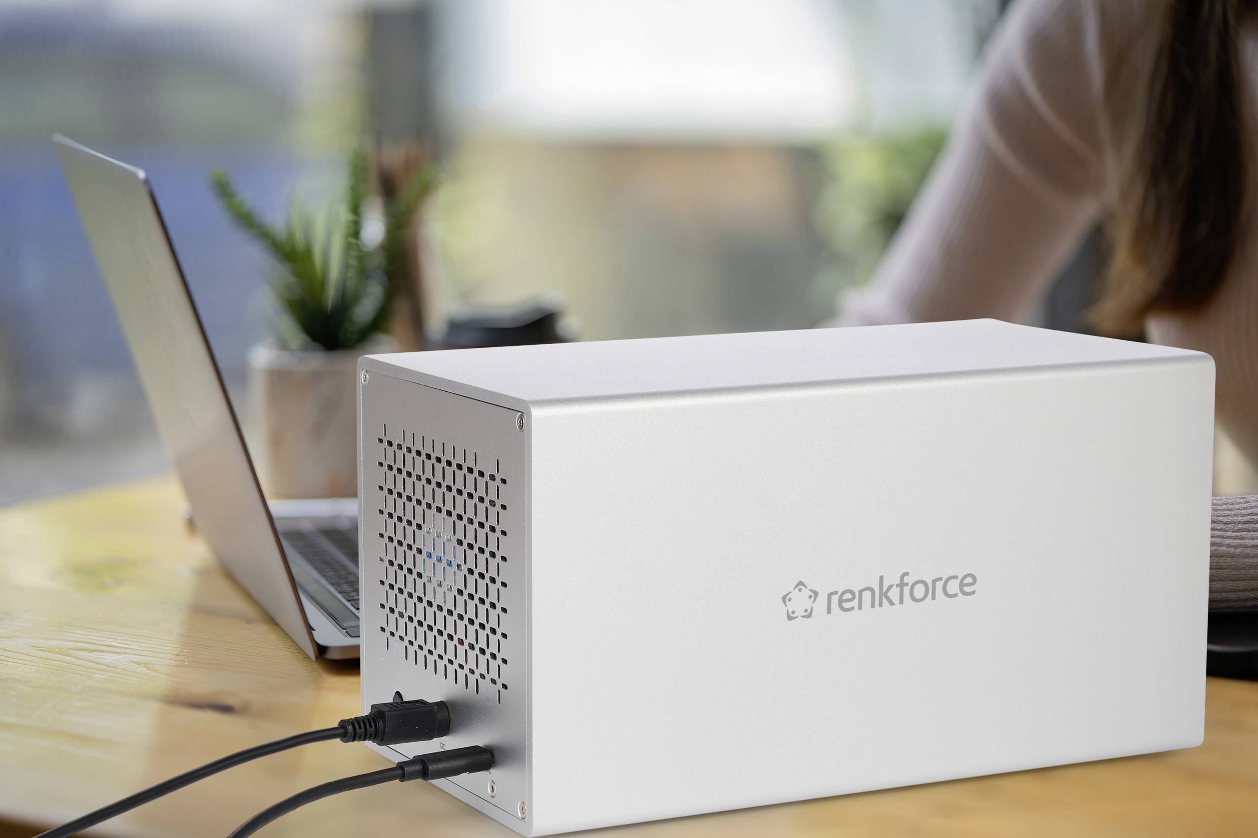 Srebrna obudowa 'renkforce' leżąca na stole obok otwartego laptopa. Do obudowy podłączonych jest kilka kabli.