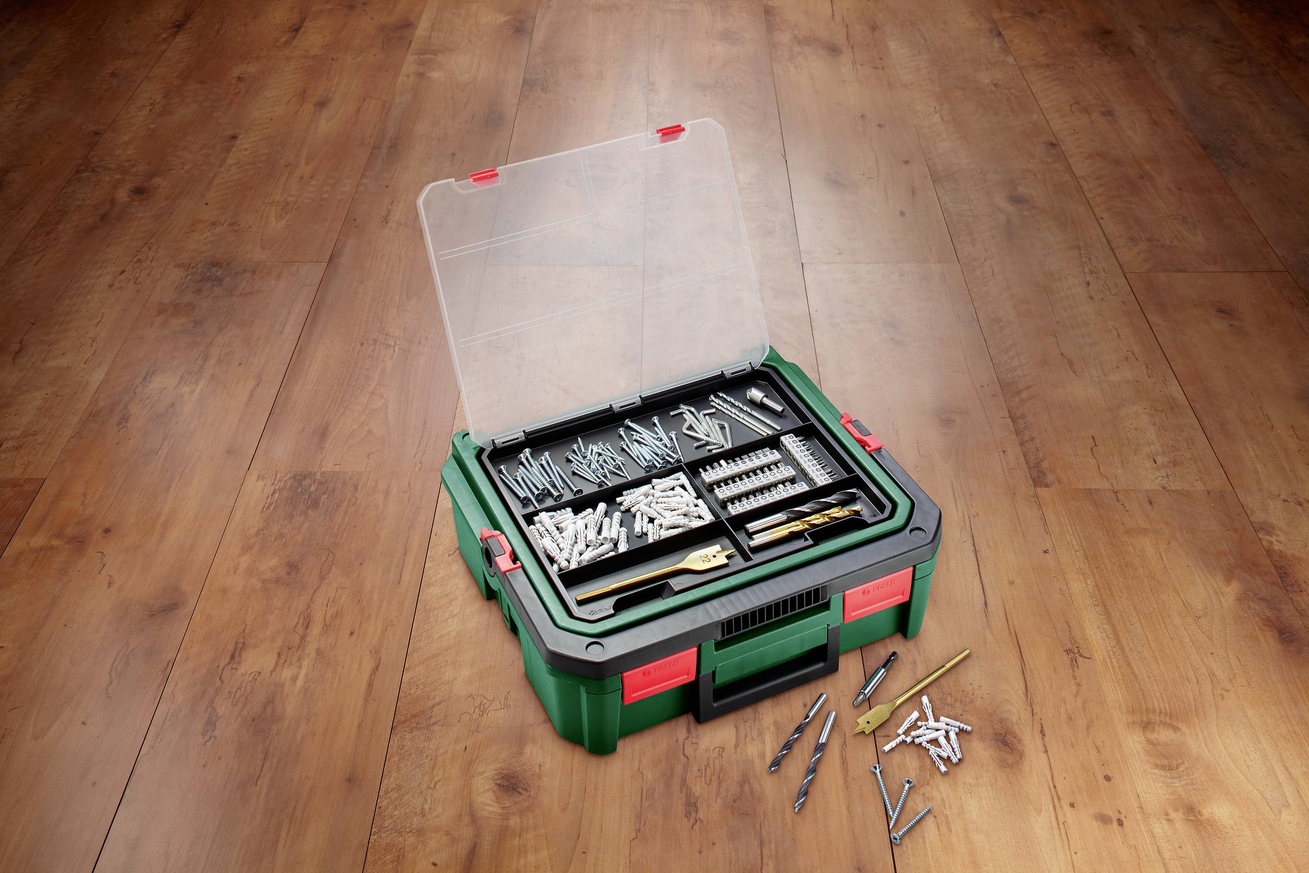 Otwarty kufer z narzędziami na drewnianej podłodze zawiera różne śruby i wiertła, posortowane w przegródkach, niektóre elementy leżą luzem obok.