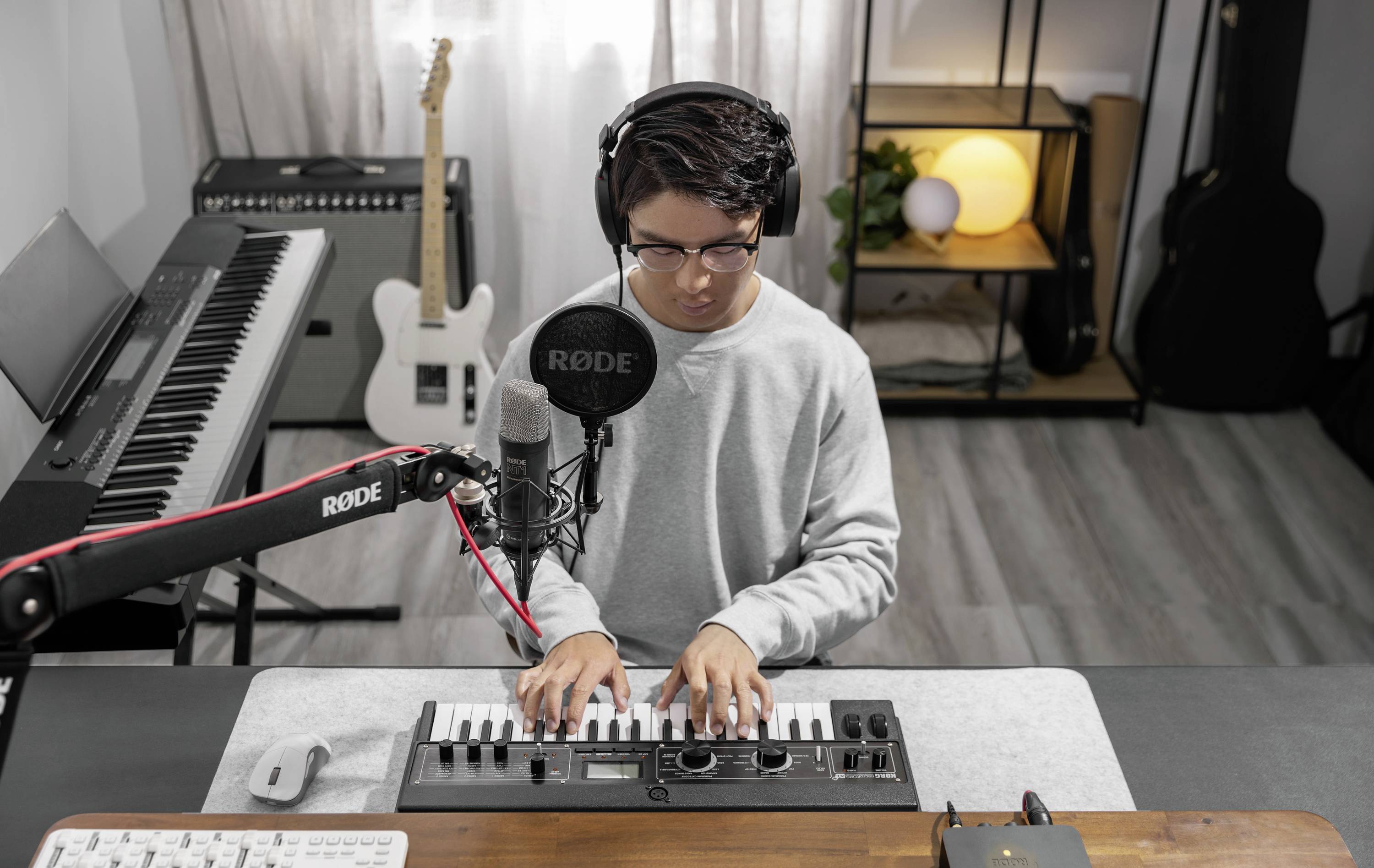 Osoba gra na małej elektronicznej klawiaturze, ma słuchawki na uszach i siedzi przed mikrofonem w studiu muzycznym.