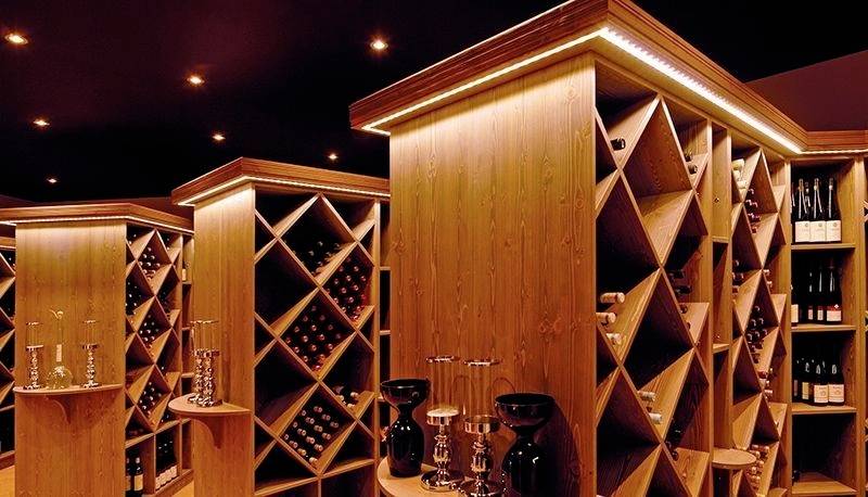 Pomieszczenie z podświetlonym piwniczką winiarską: drewniane ściany i regały z ukośnymi przegrodami, wypełnione butelkami wina i kieliszkami do wina.