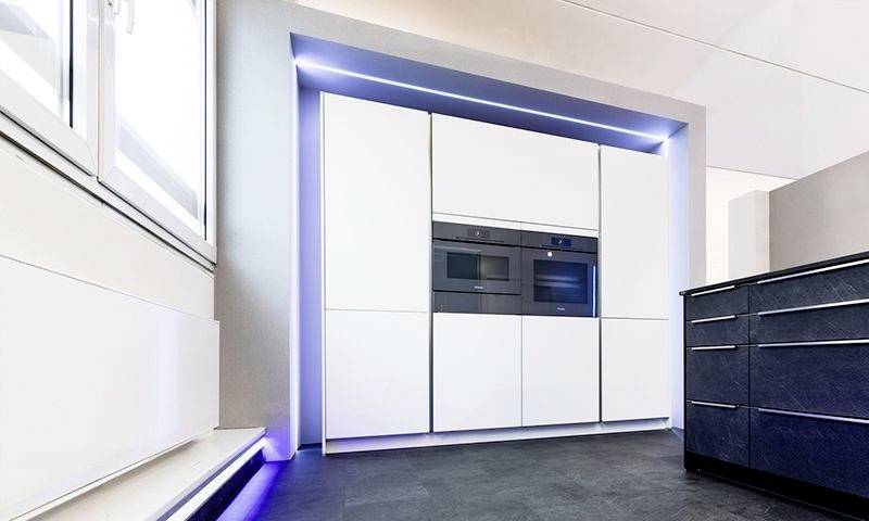Nowoczesna kuchnia z białymi szafkami zabudowanymi, zintegrowanymi urządzeniami i ciemną wyspą kuchenną. Minimalistyczny design, dużo oświetlenia.
